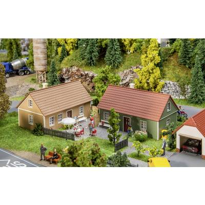 Petit village miniature avec deux maisons, des personnages assis et marchant, des arbres en arrière-plan, représentation d'une scène rurale.