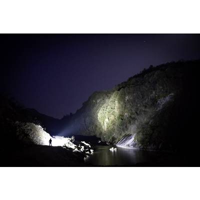 Une personne se tient au bord de la rivière par une nuit sombre, une lampe de poche éclairant les alentours. Des collines et de l'eau entourent la scène.