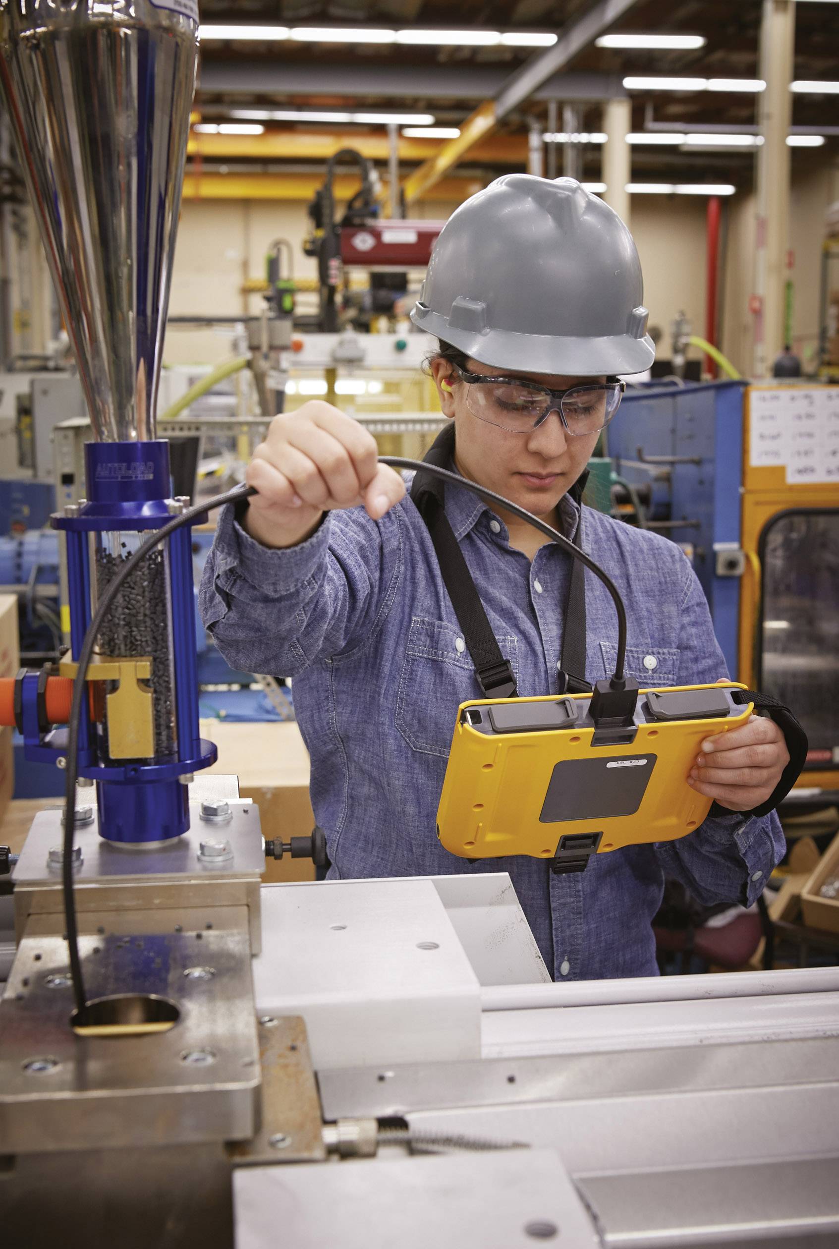 Une personne portant un casque de protection et des lunettes travaille avec un appareil numérique dans un environnement industriel, entourée de machines.