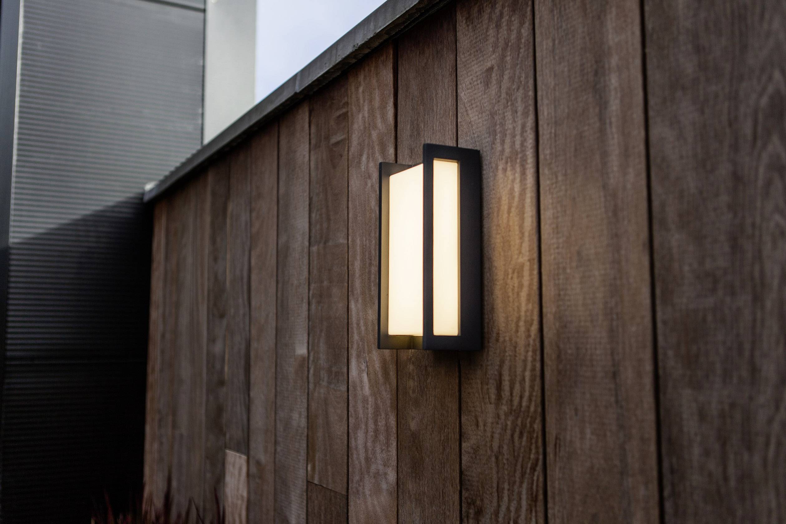 Applique extérieure illuminant un mur en bois moderne, éclairant doucement les alentours. Idéal pour une ambiance extérieure chaleureuse et élégante.
