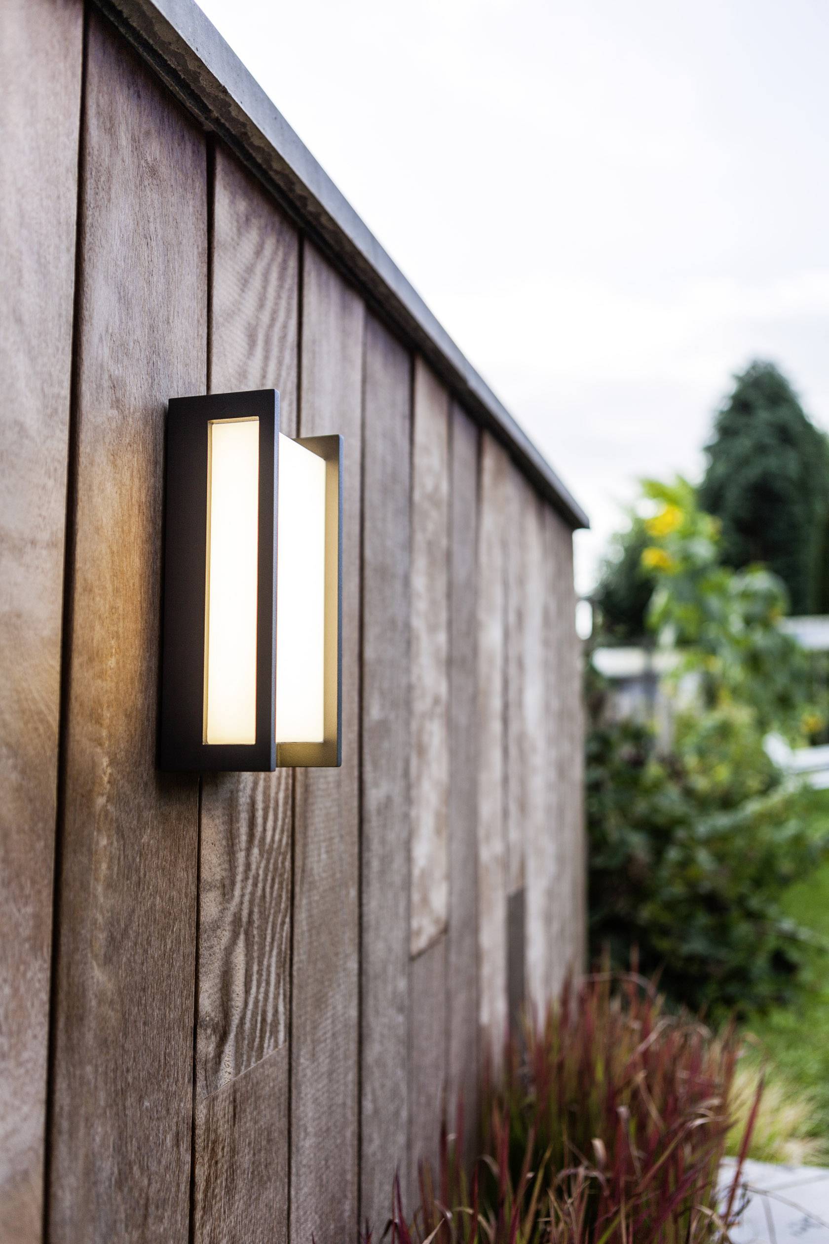 Lampe extérieure sur un mur en lattes de bois dans un jardin, allumée au crépuscule. Des plantes sont visibles en arrière-plan.