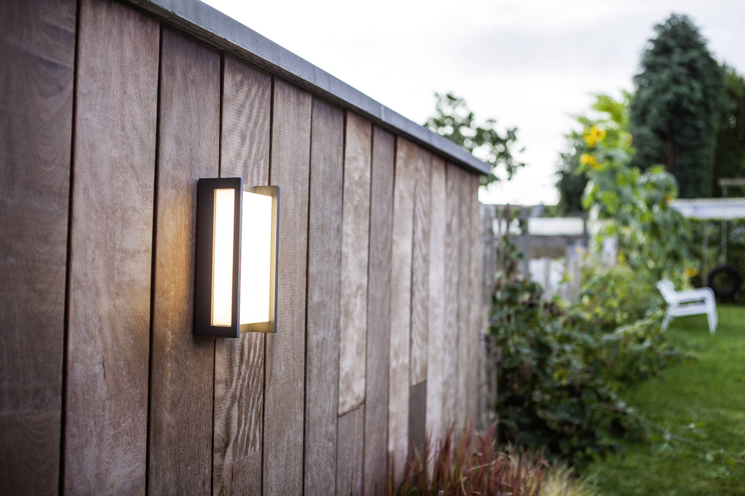 Clôture en bois avec un luminaire extérieur moderne, brillant au crépuscule ; un jardin avec des arbres et des plantes en arrière-plan.