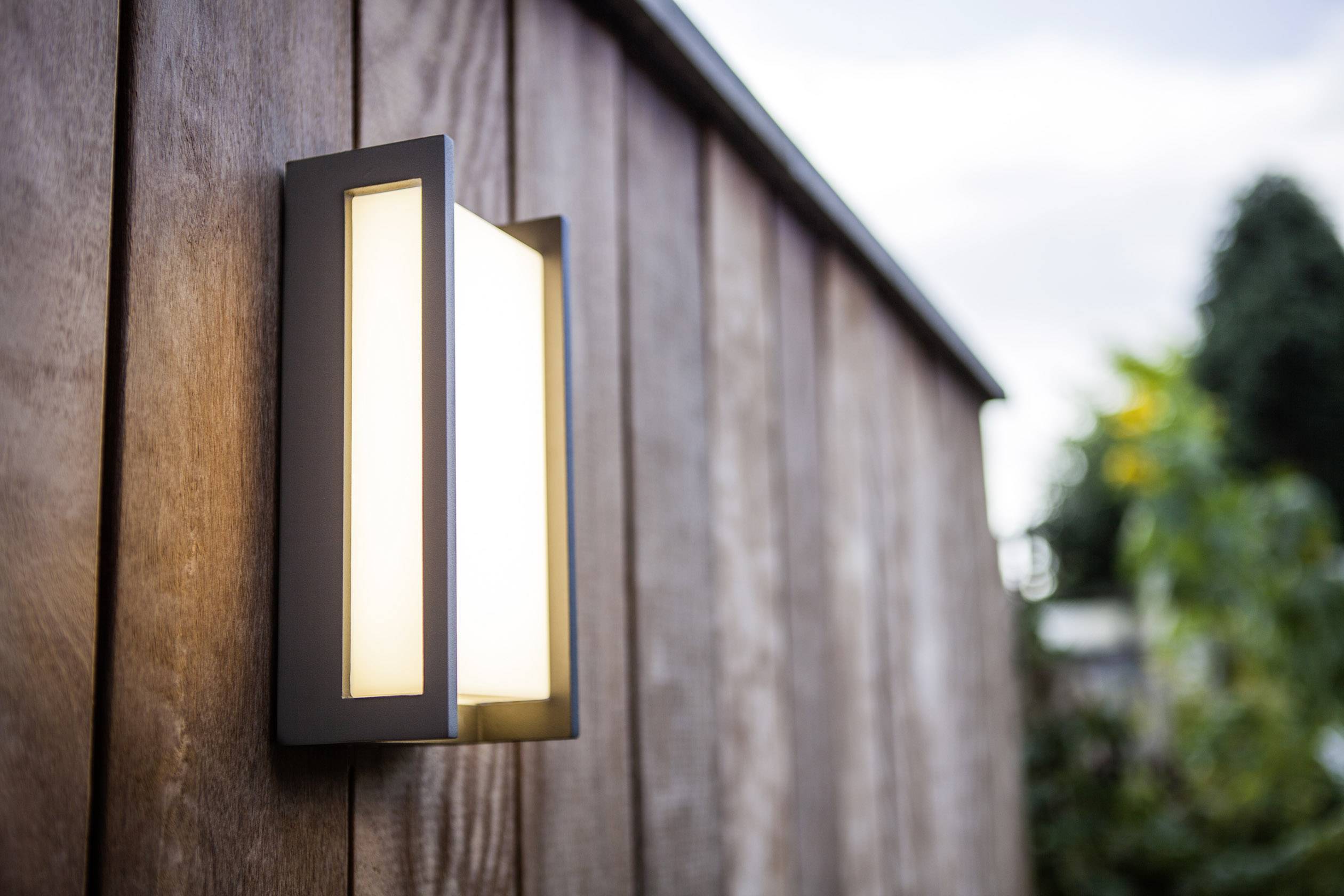 Un luminaire moderne est fixé à un mur en bois et éclaire les alentours au crépuscule.