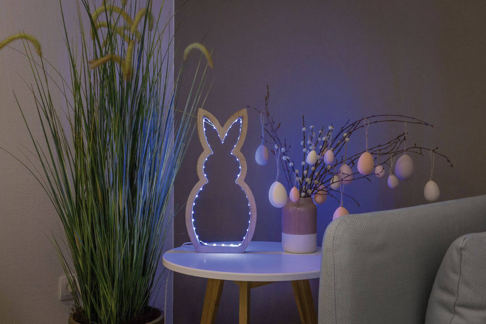 Sur une petite table se trouve une figurine de lapin lumineuse et un vase contenant des branches avec des œufs de Pâques suspendus. À gauche, on peut voir une plante haute.