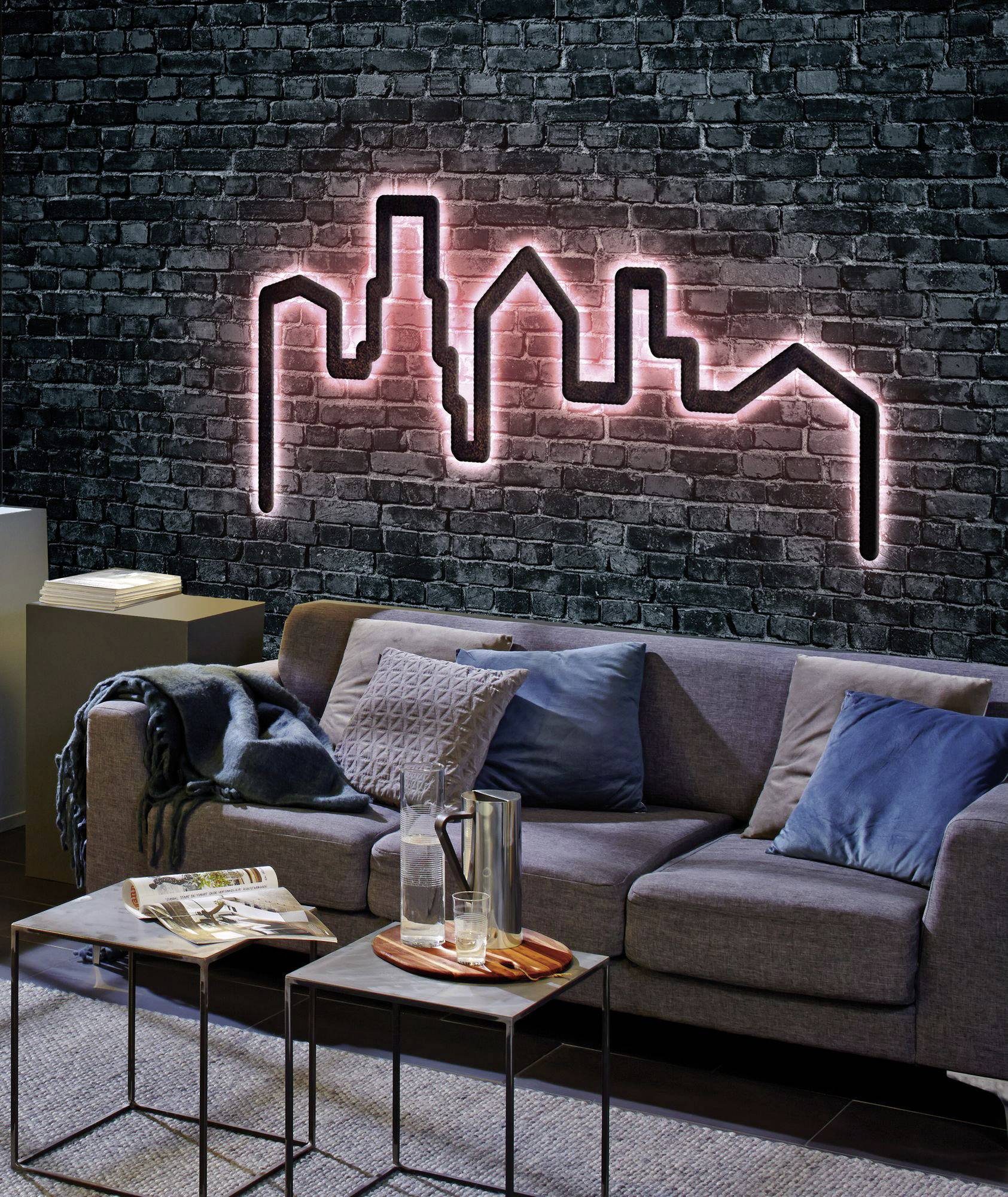 Salon avec un canapé gris, des coussins bleus et un plaid. Sur le mur de briques, un panorama urbain néon rose s'illumine.