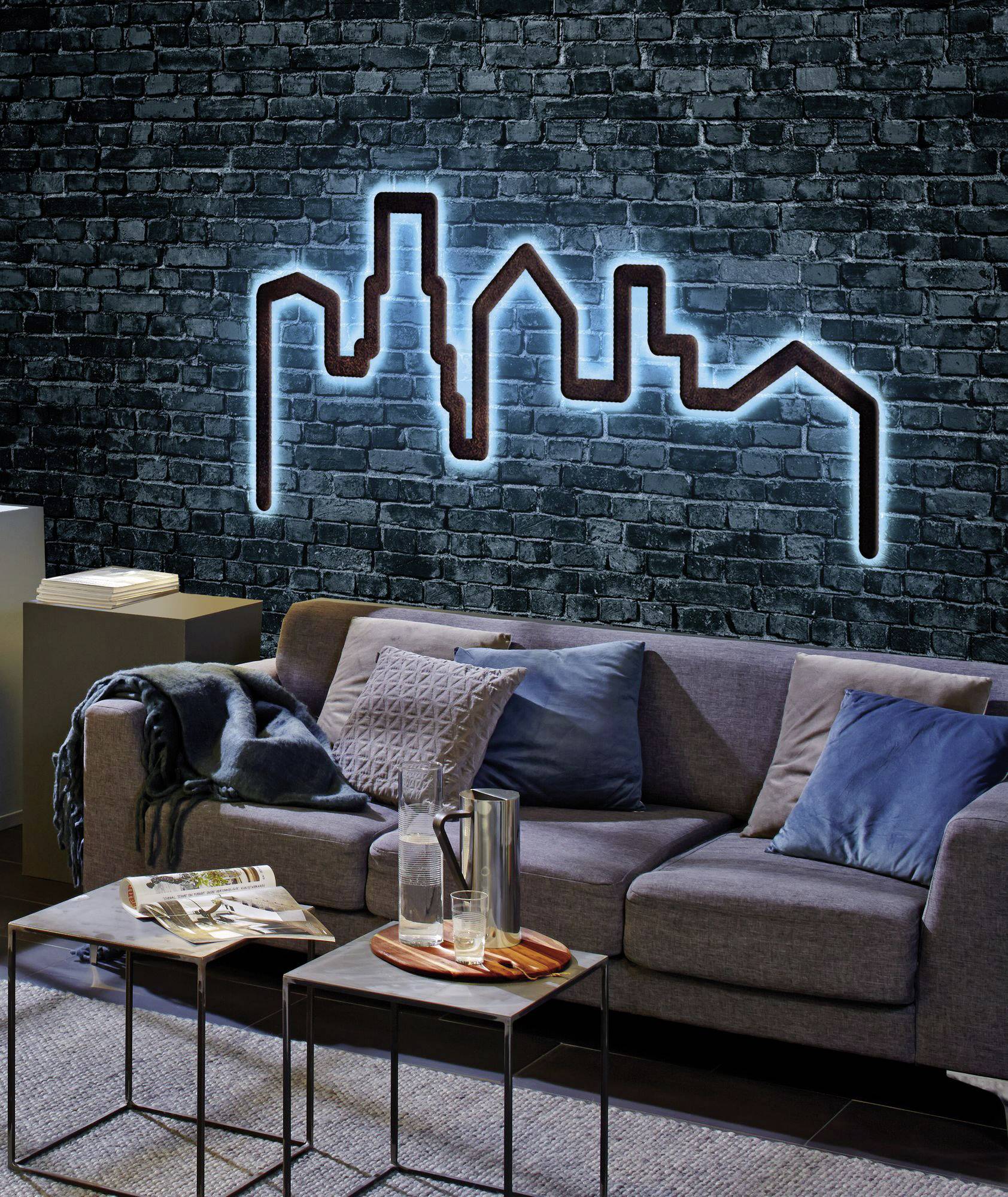 Un salon moderne avec un canapé gris, décoré de coussins bleus et gris. Sur le mur, un tableau de ville lumineux en néon.