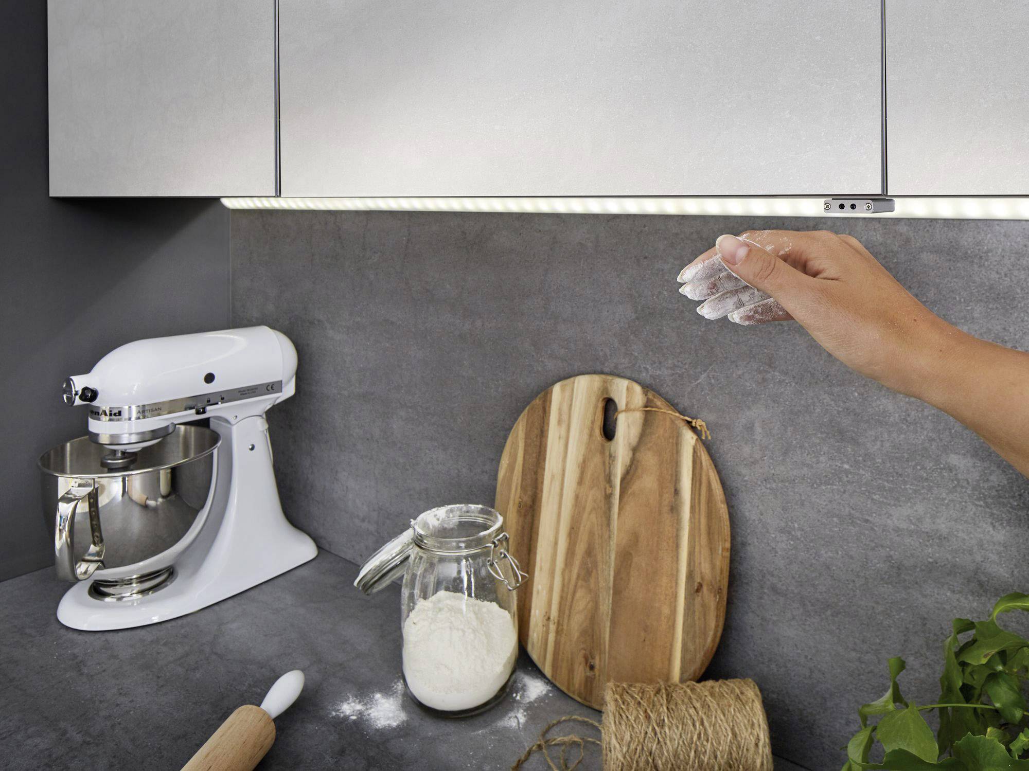 Une main saupoudre de la farine sur un plan de travail dans une cuisine moderne. Un mixeur, un verre de farine et des planches en bois sont visibles.