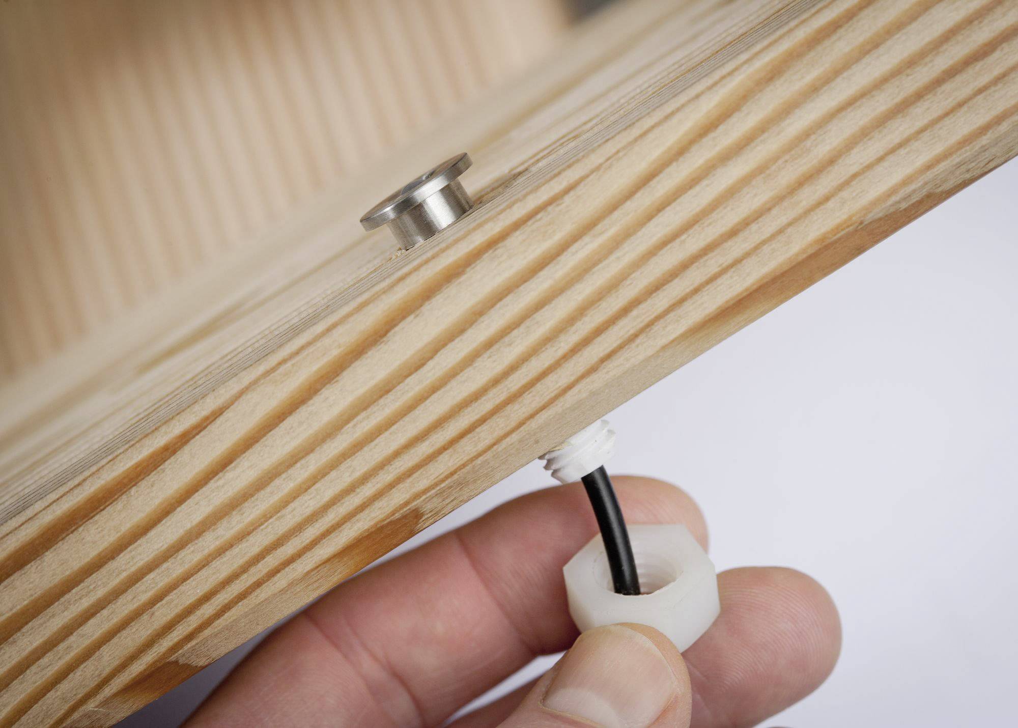 Gros plan d'une planche en bois avec une vis métallique et une main guidant un fil noir à travers un écrou en plastique blanc.