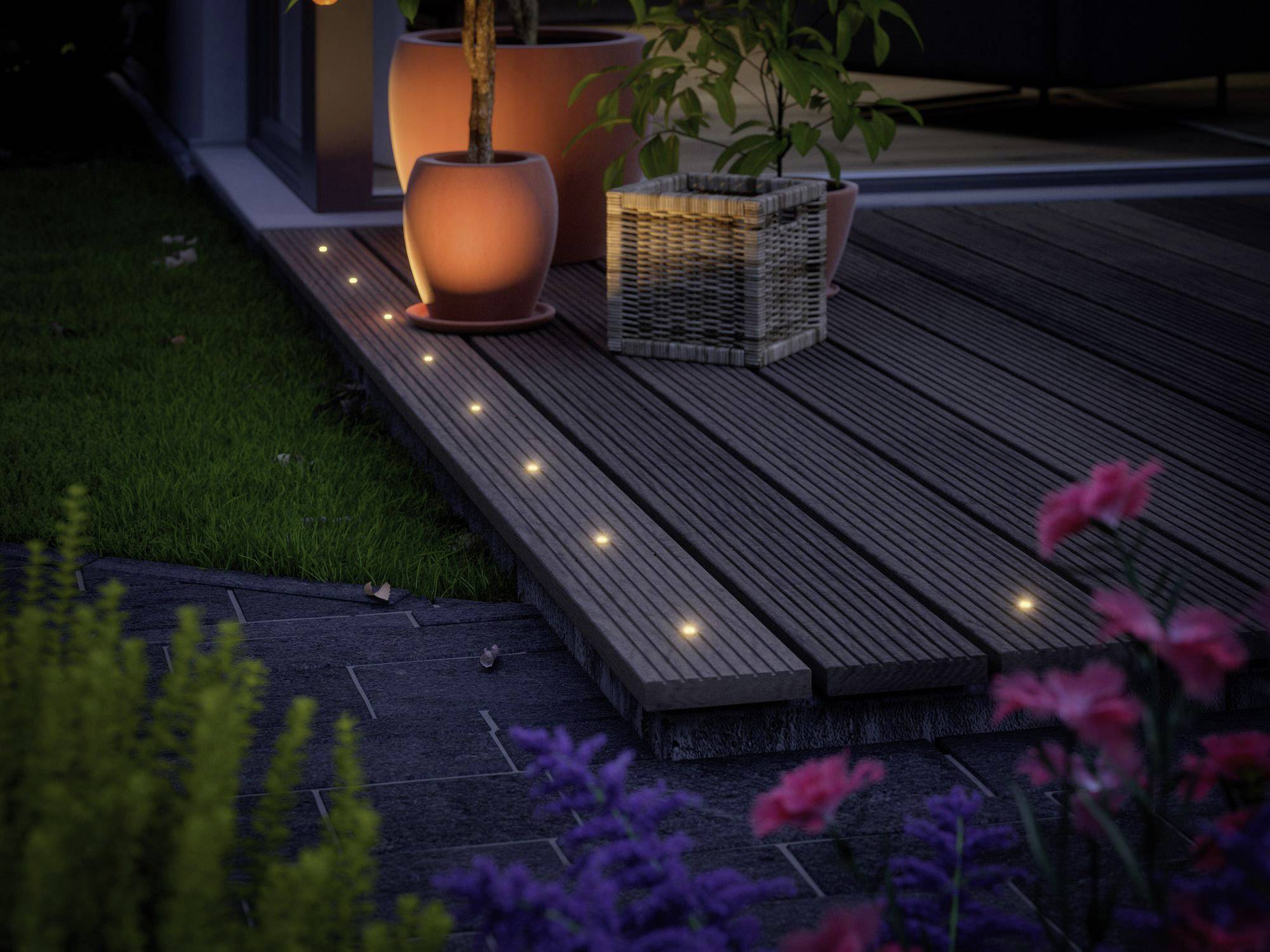 Terrasse en planches de bois, entourée de pots de fleurs illuminés et de plantes de jardin. De petites lumières sont encastrées dans le sol.