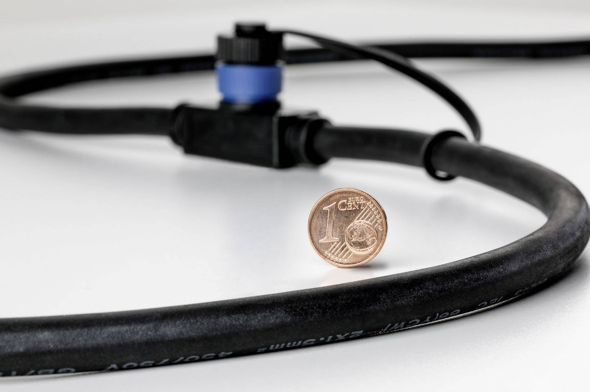 Un tuyau d'irrigation noir est posé sur une table, à côté d'une pièce de monnaie de un centime. Le tuyau est équipé d'un embout de valve bleu.