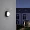 Luminaire extérieur sur un mur gris éclairant une cour avec des bâtiments modernes en arrière-plan au crépuscule.