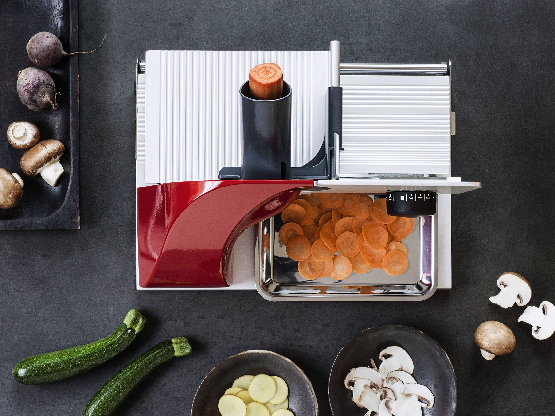 Une mandoline avec un élément rouge coupe des rondelles de carottes. Sur la table sont posés des courgettes, des champignons, des betteraves et des pommes de terre coupées.