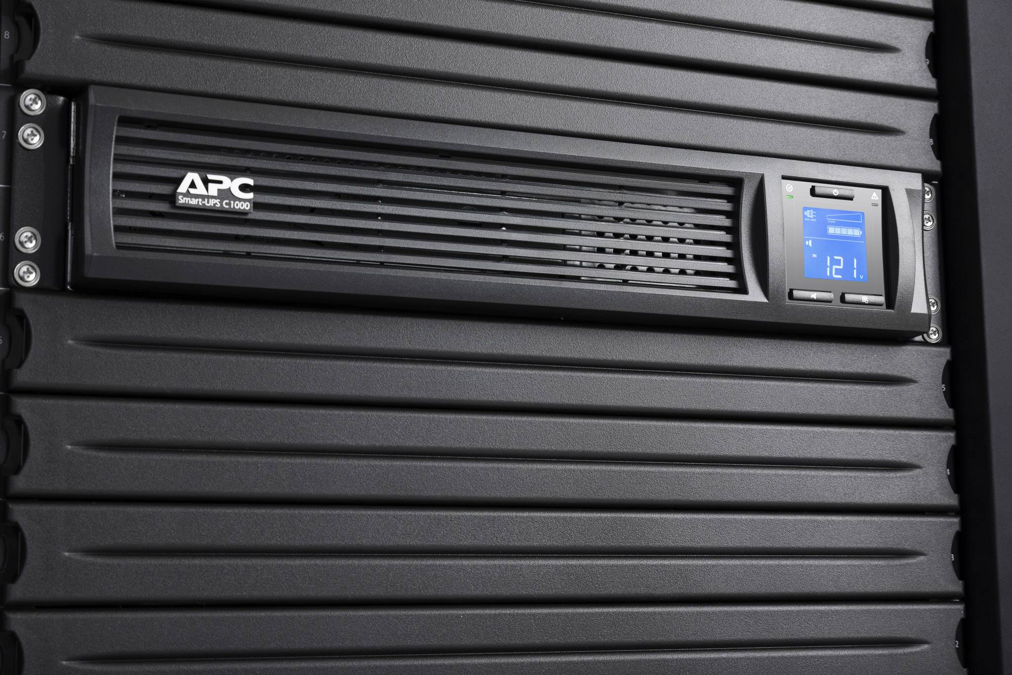 Un onduleur APC Smart-UPS à l'avant d'un rack serveur noir. L'écran de l'onduleur affiche une tension de sortie de 121 V.