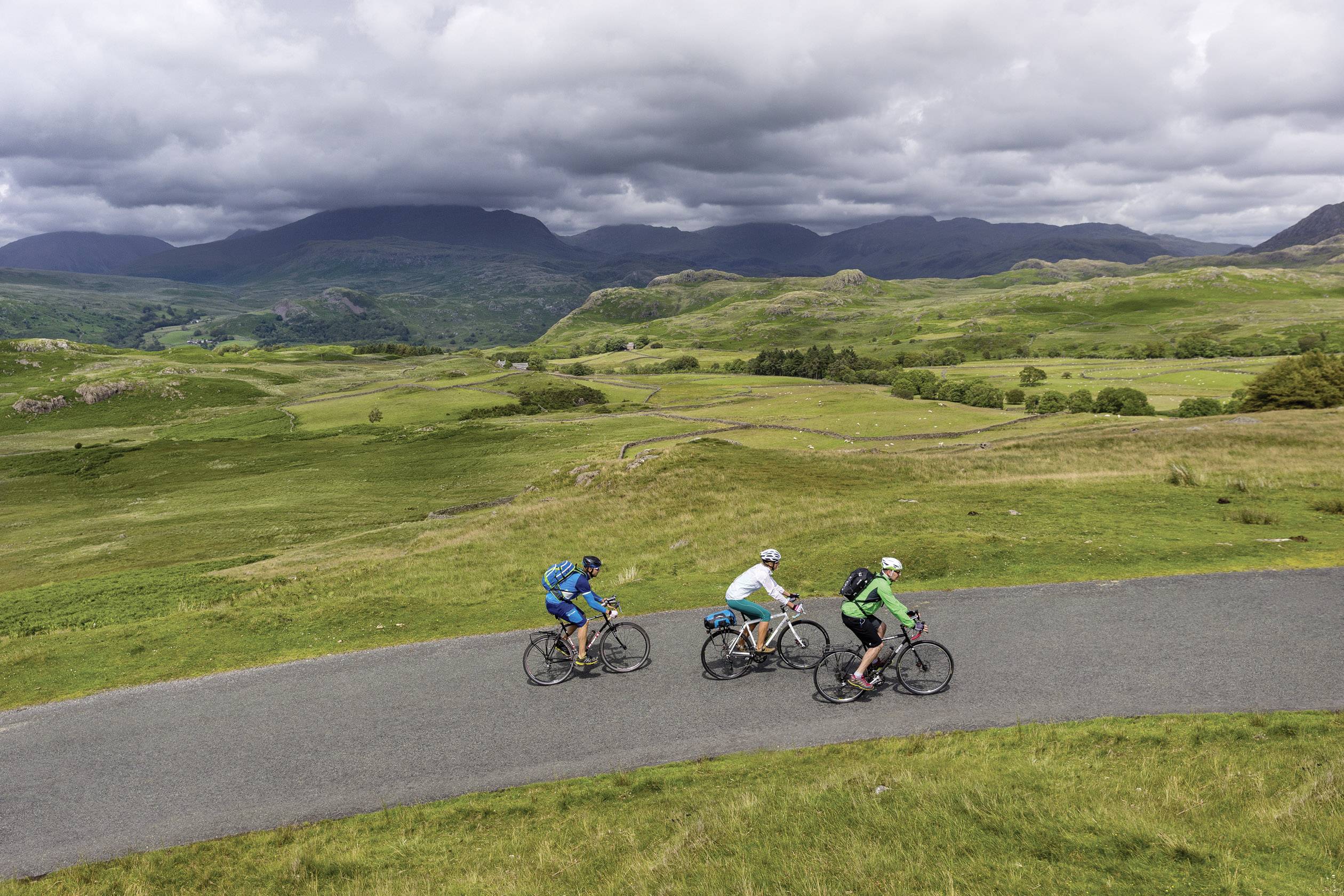 Trois cyclistes roulent sur une route à travers un paysage vallonné et verdoyant, sous un ciel nuageux.