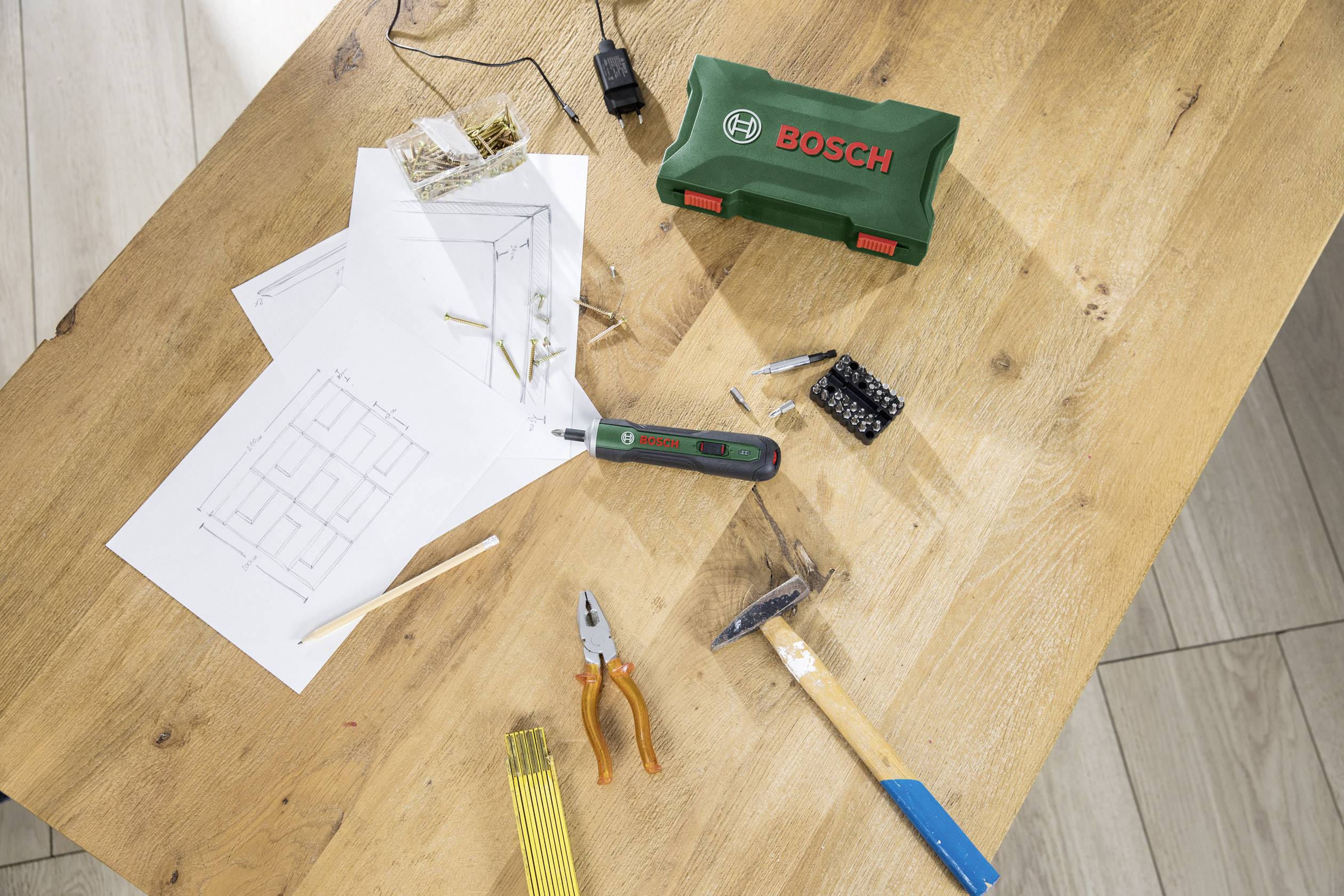 Une surface de travail avec des outils : des plans, des vis, une visseuse, une pince, un marteau et un coffret à outils vert portant le logo 'BOSCH'.