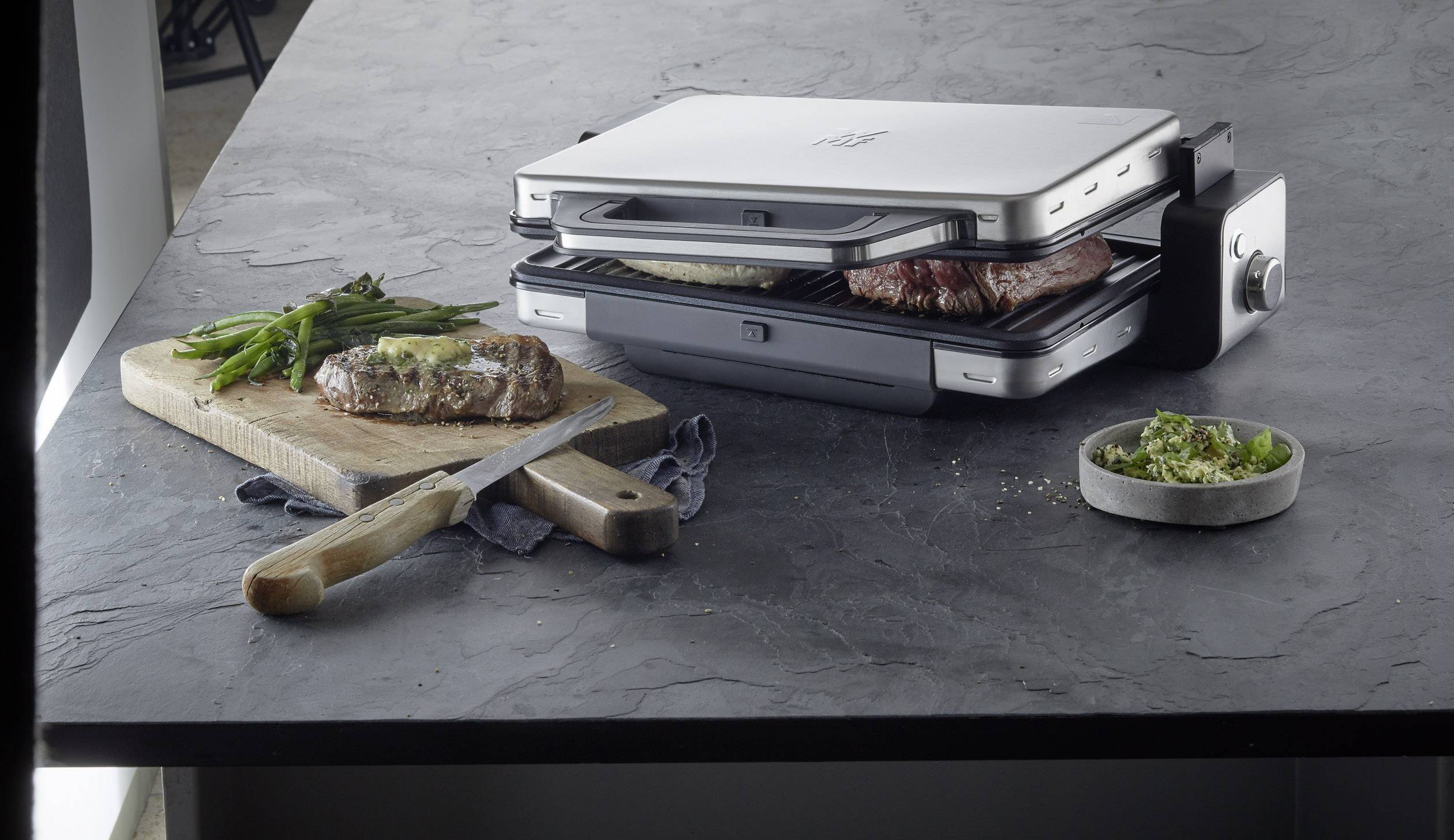 Un grill de contact en acier inoxydable avec des plaques ouvertes fait griller deux morceaux de viande. À côté, une planche en bois présente un steak prêt à être servi et des légumes.