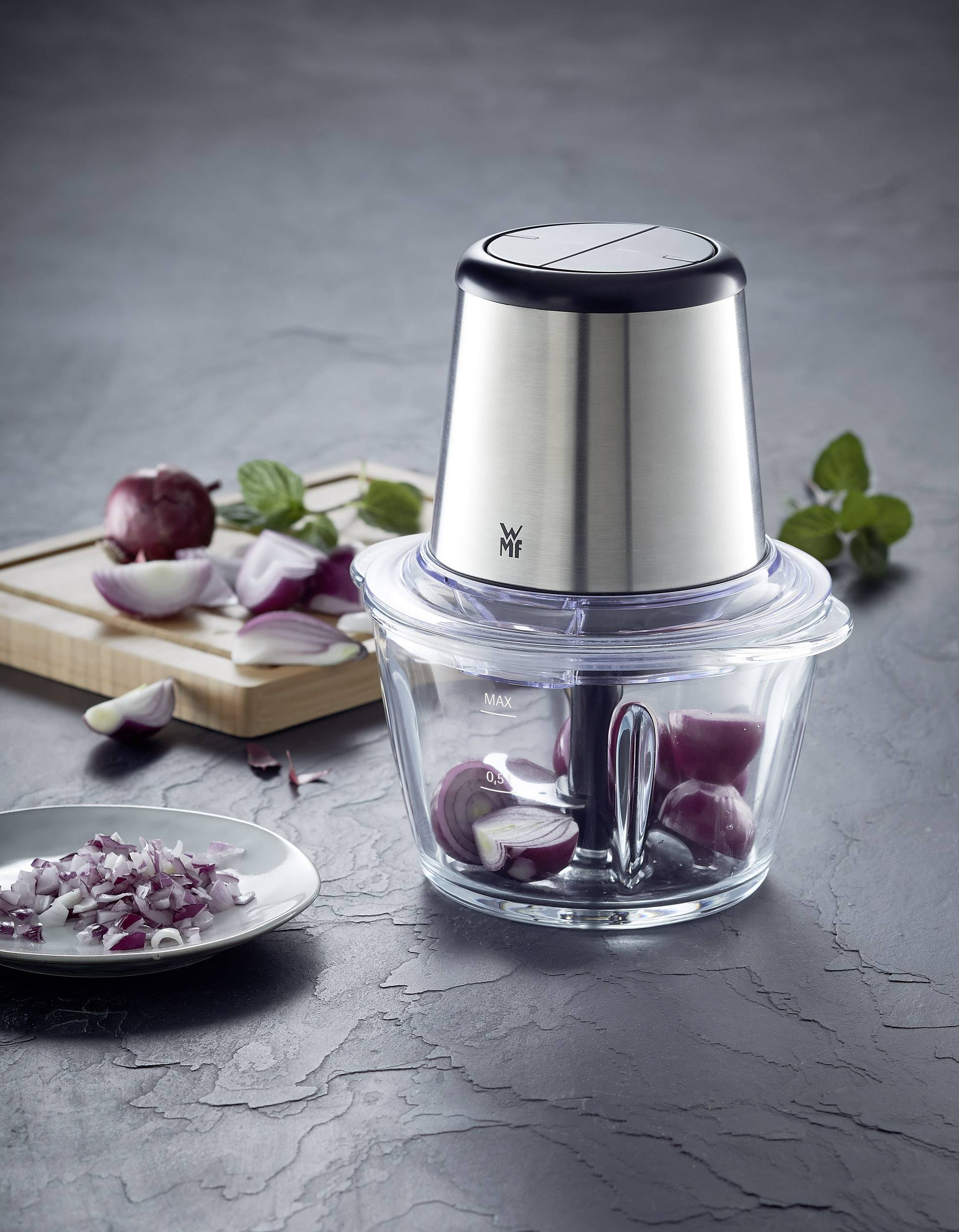 Un hachoir électrique avec un accessoire en acier inoxydable est posé sur un plan de travail de cuisine ; à côté se trouve une planche à découper en bois avec des morceaux d'oignon.