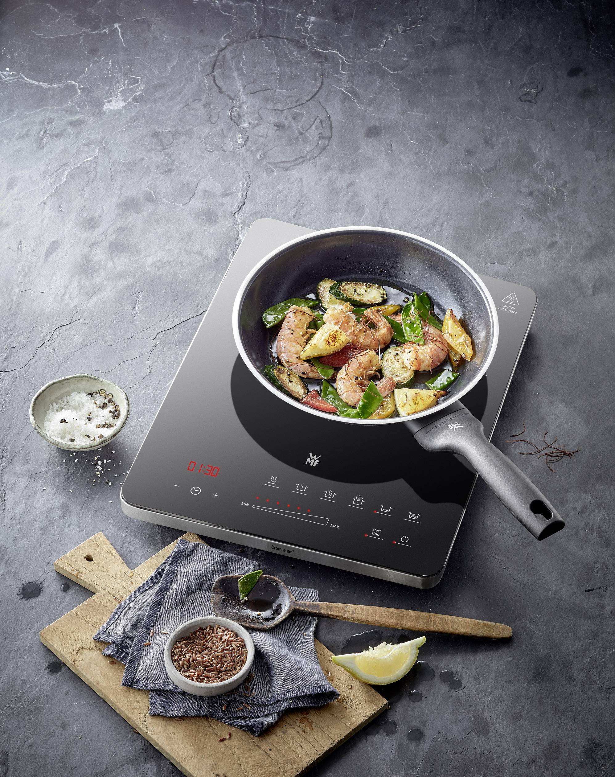 Une poêle avec des légumes et des morceaux de viande sur une plaque à induction. À côté, des bols d'épices et un morceau de citron sont disposés.