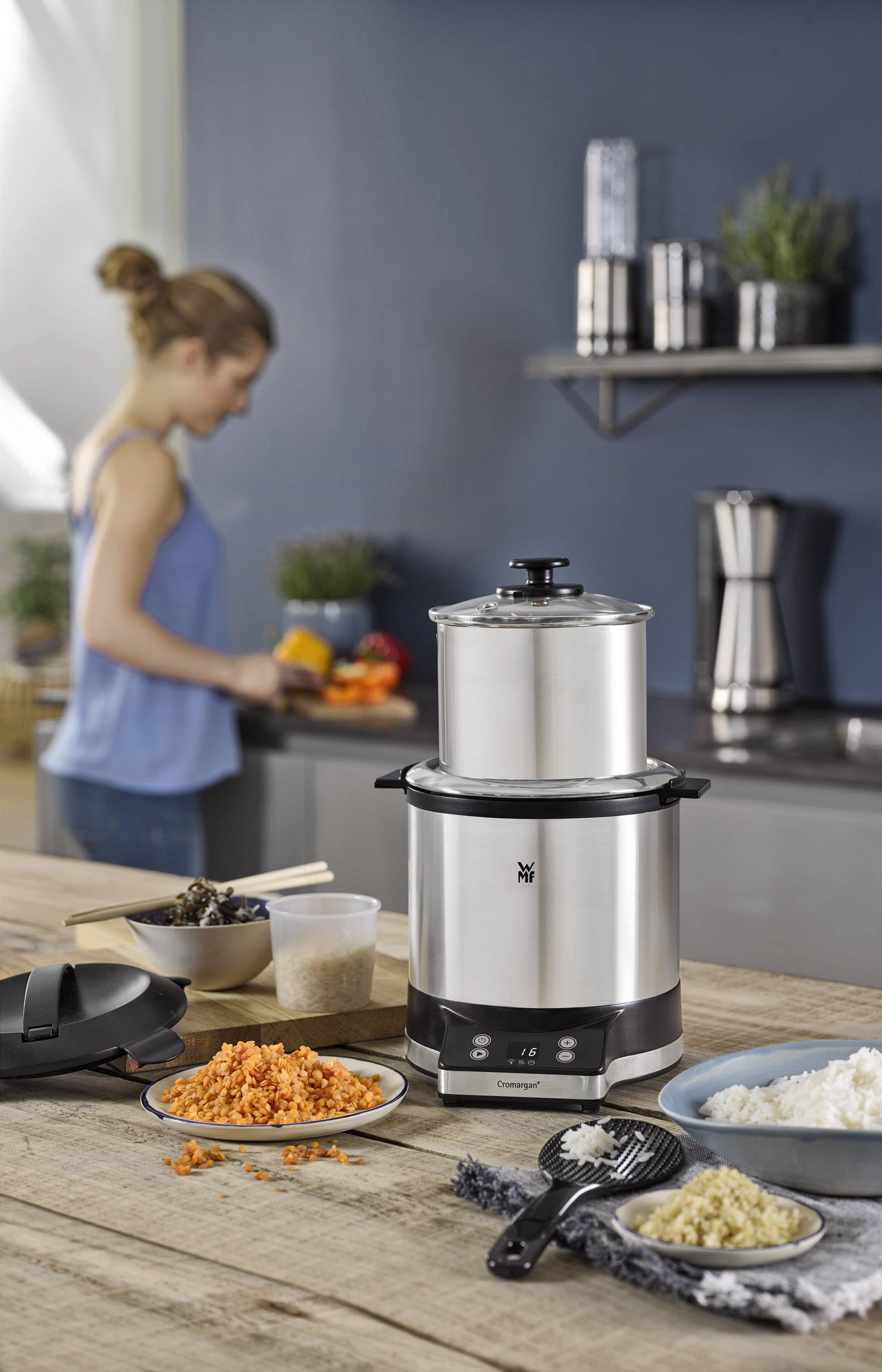 Cuiseur vapeur sur une table en bois, à côté des légumes coupés et du riz. En arrière-plan, une personne cuisinant dans une cuisine moderne.