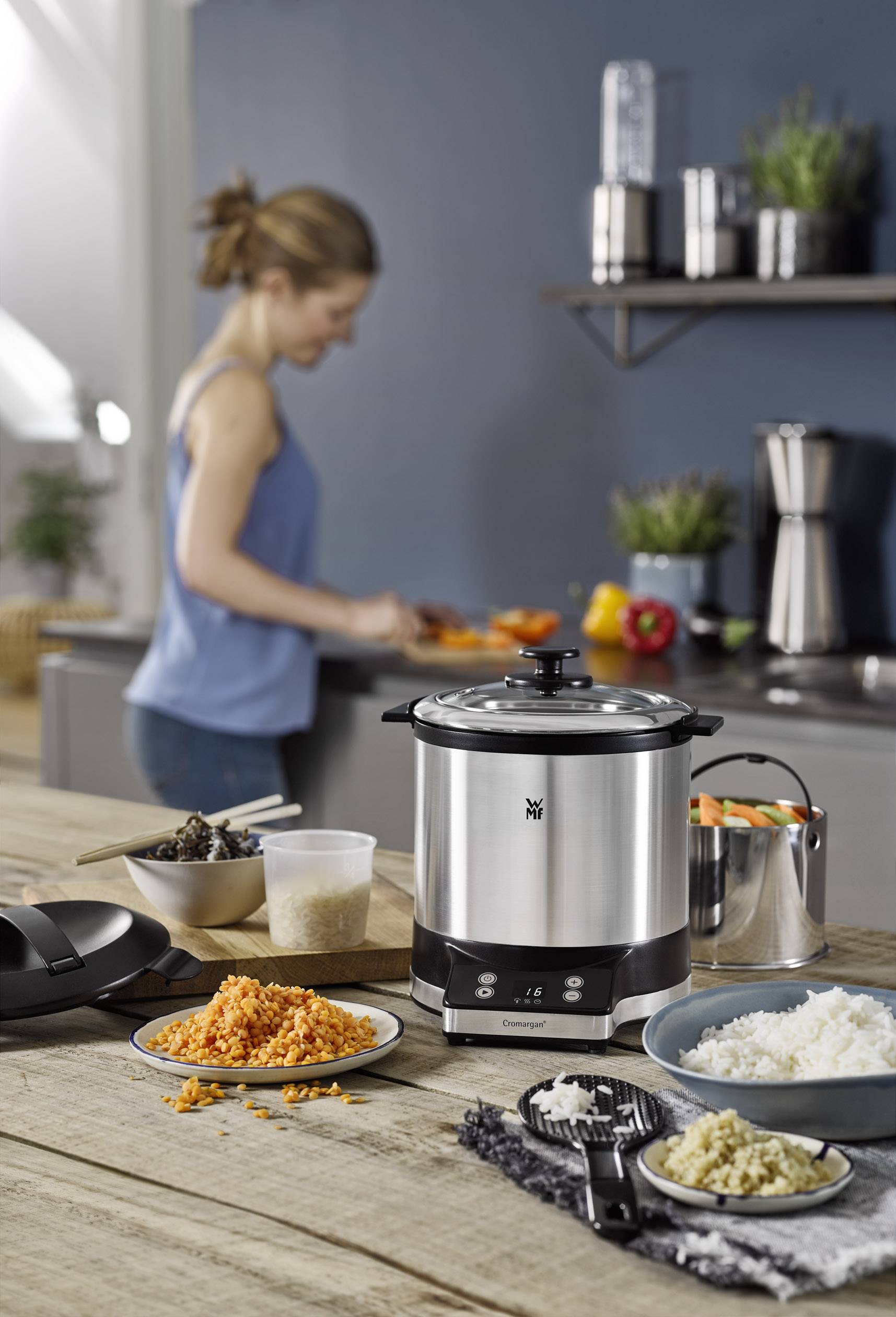 Une femme coupe des légumes dans une cuisine moderne. Au premier plan, un cuiseur à riz électrique est posé avec des ingrédients tels que des lentilles et du riz.