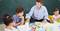 Un homme est assis à une table avec trois enfants et joue à un jeu de plateau éducatif. Un tableau vert est en arrière-plan.