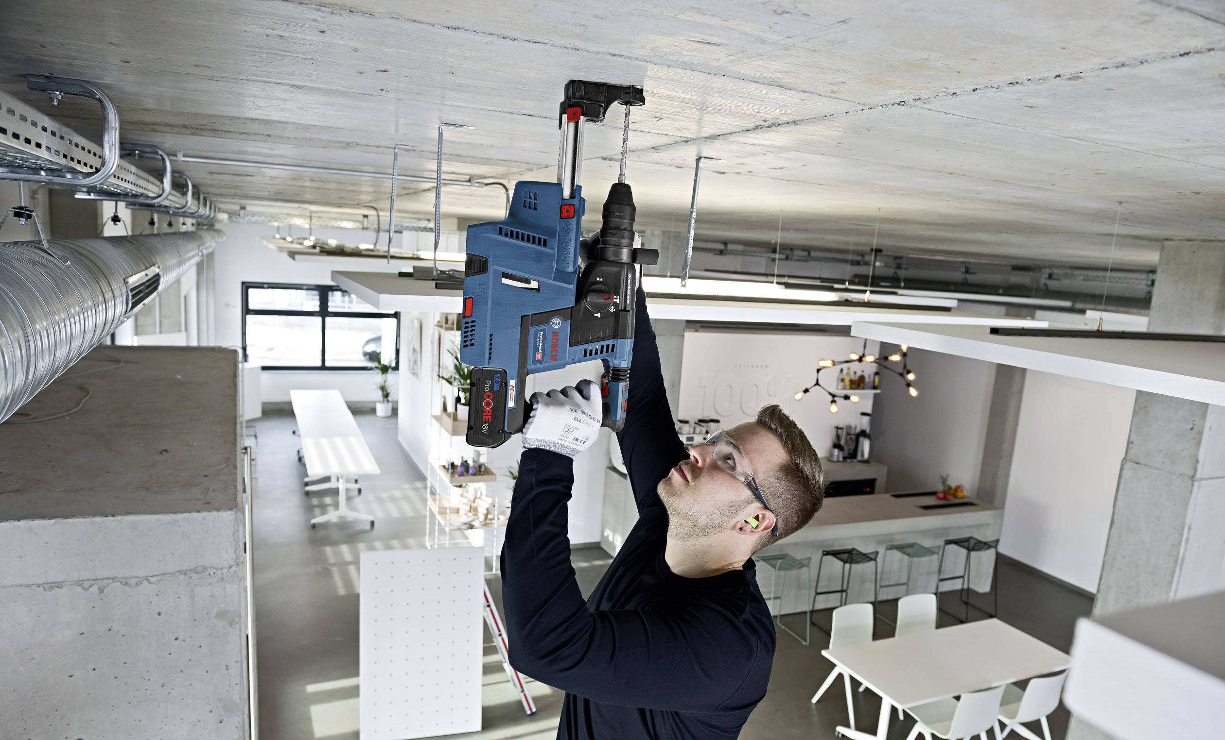 Un homme fore un plafond avec une grande perceuse électrique dans un bâtiment de bureau moderne. Il porte des lunettes de protection et des gants.