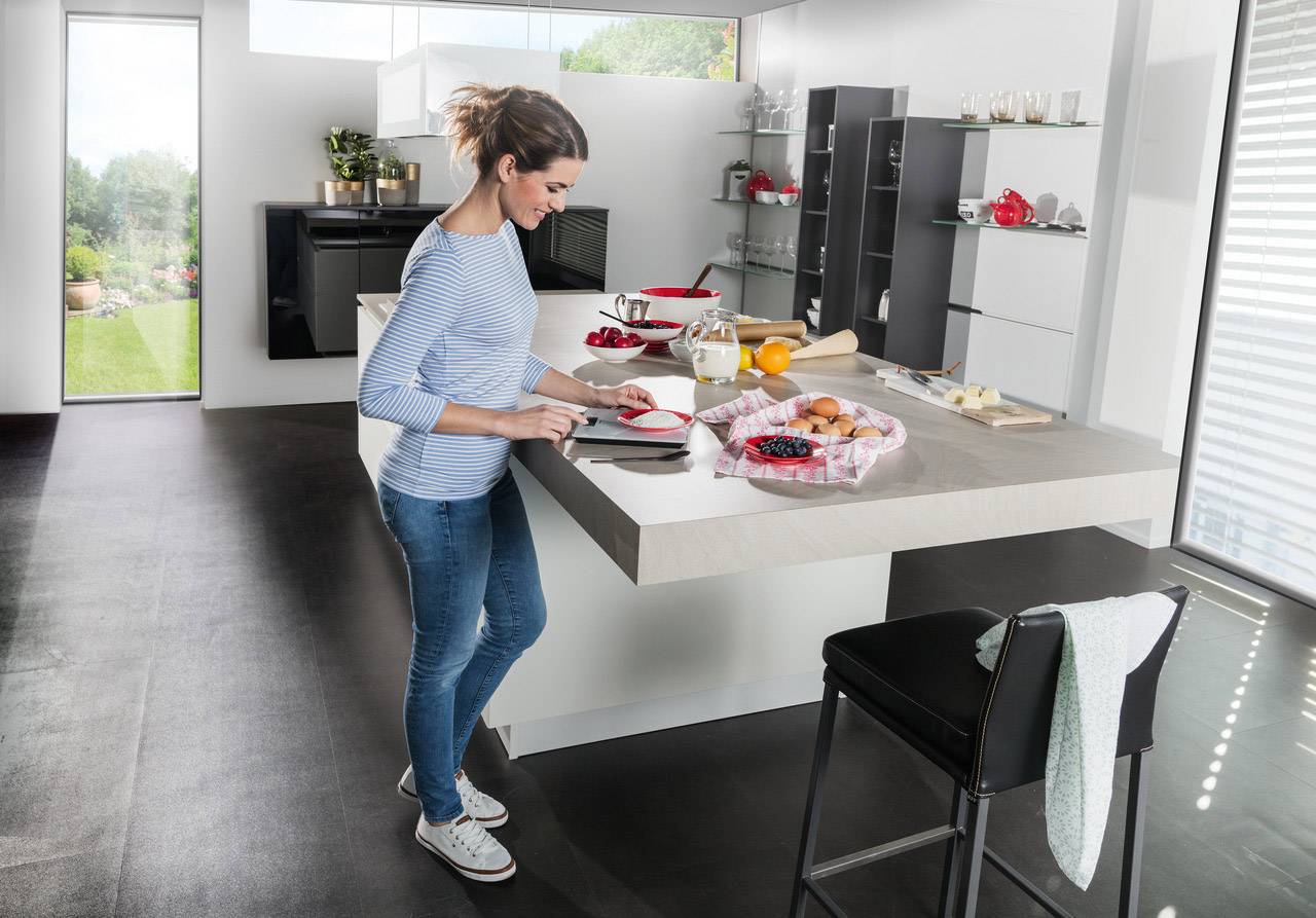 Une femme en jeans et pull rayé coupe des fruits dans une cuisine moderne. Une assiette avec des œufs et un verre de jus sont posés à côté.