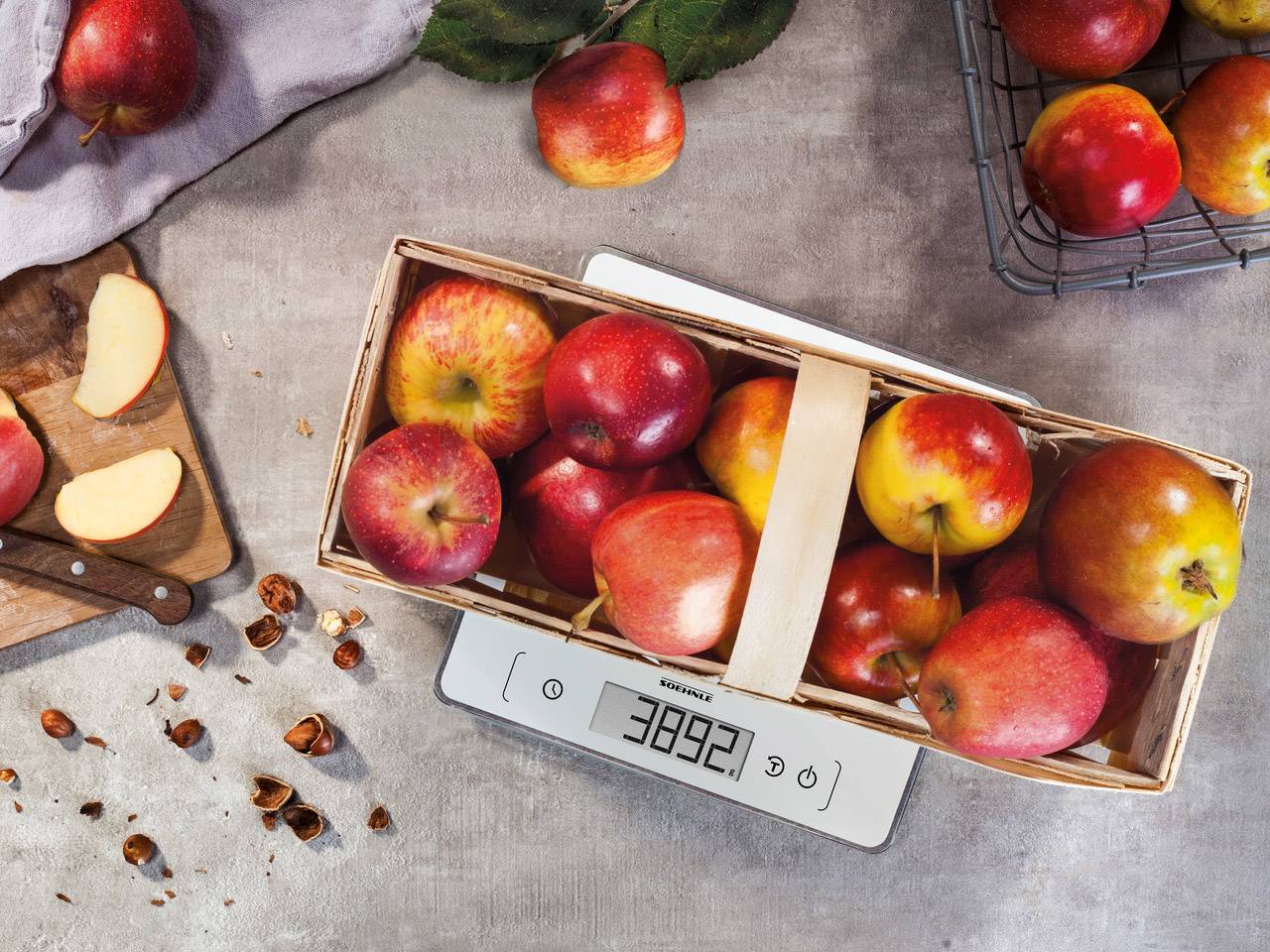Un panier rempli de pommes rouges est posé sur une balance numérique affichant '3892 g'. À côté de la balance, il y a des tranches de pommes et des noisettes.