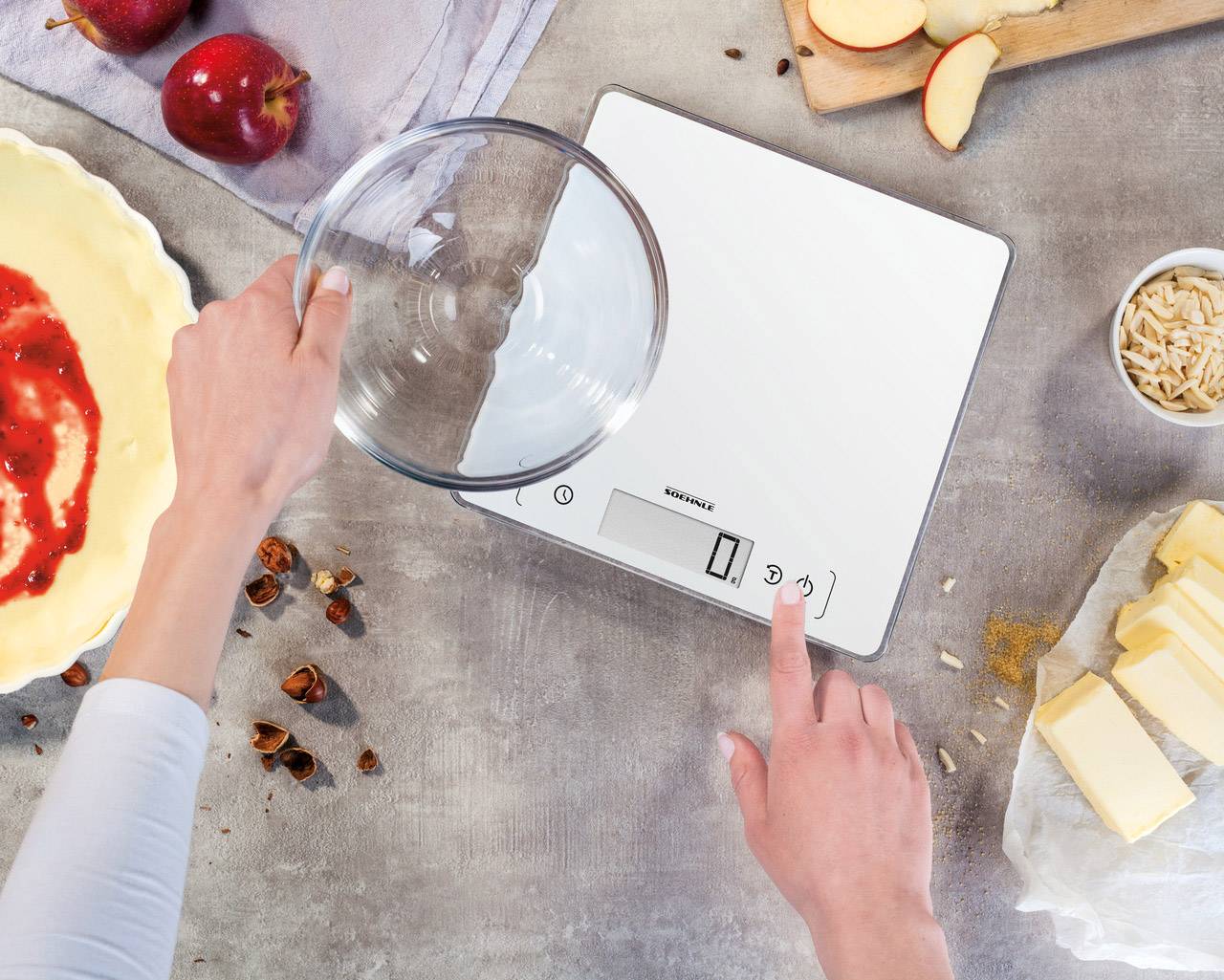 Une personne pèse des ingrédients dans un bol en verre sur une balance de cuisine numérique, entourée de pommes, de pâte, de beurre et d'amandes.