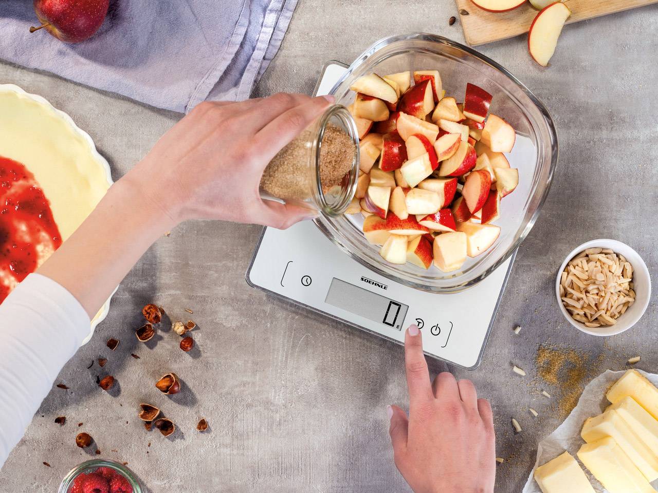 Une personne pèse un bol de pommes coupées sur une balance de cuisine numérique. À côté, on trouve des ingrédients tels que du beurre et des amandes.