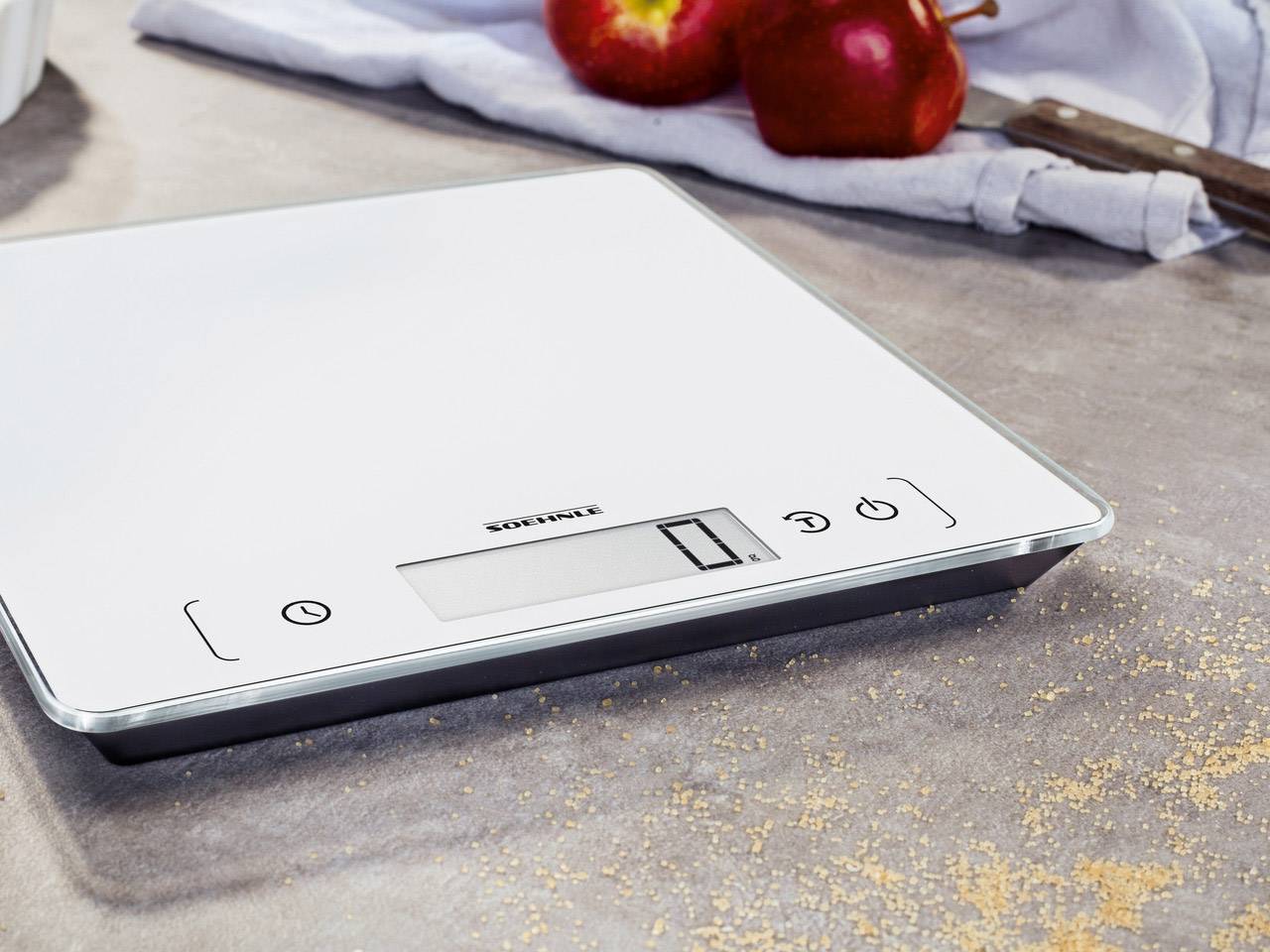 Balance de cuisine électronique sur une table de cuisine avec des pommes en arrière-plan. Affichage LCD et touches de commande visibles. Idéale pour peser des ingrédients.