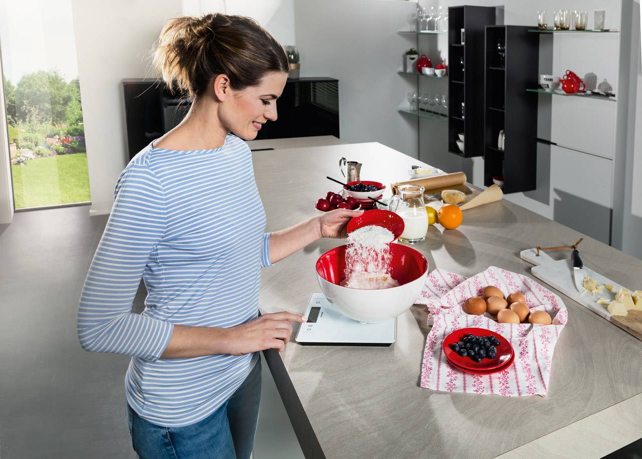 Une femme dans un espace de cuisine moderne pèse de la farine dans un bol rouge et blanc sur une balance de cuisine, entourée d'ingrédients.