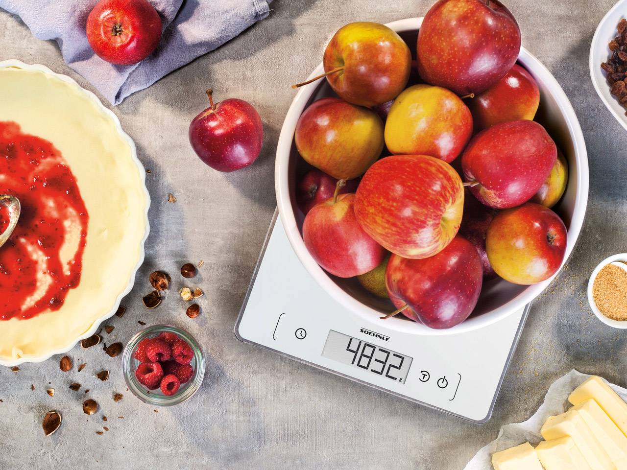 'Un bol de pommes rouges sur une balance de cuisine numérique affichant 4832, entouré d'ingrédients de pâtisserie tels que des framboises et du beurre.'