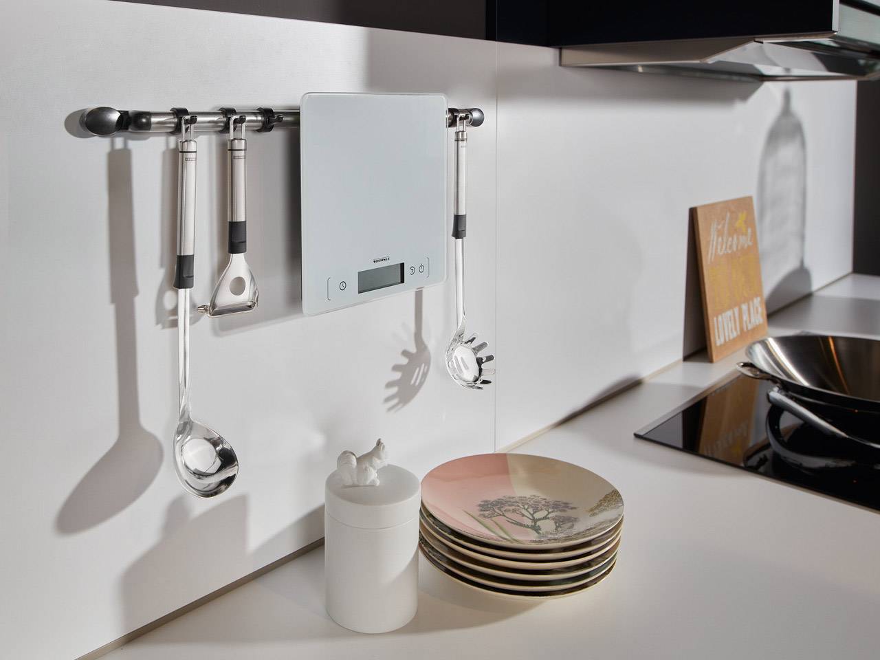 Plan de travail de cuisine avec support mural pour ustensiles et balance numérique, à côté d'une pile d'assiettes et de contenants de rangement.