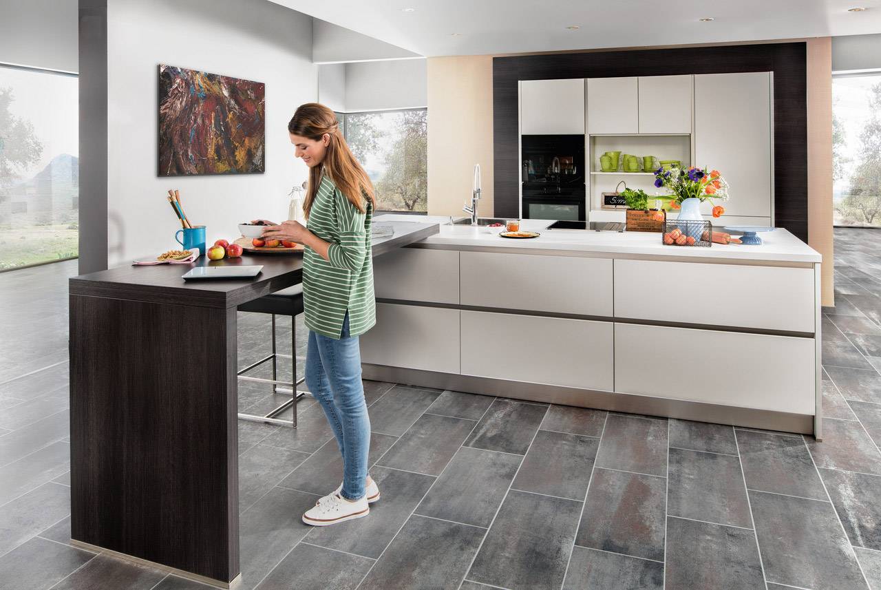 Une femme se tient dans une cuisine moderne, debout près d'un comptoir et écrit sur un bloc-notes. À côté d'elle, des fruits et des fleurs sont posés sur le comptoir.