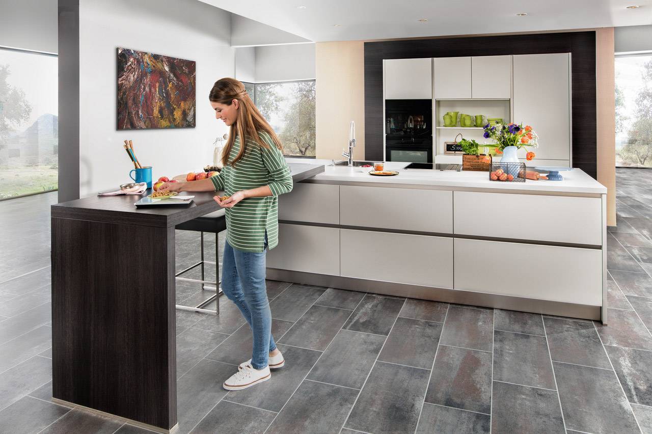 Une femme prépare un repas dans une cuisine moderne. Elle est debout à un comptoir avec un bol et des ustensiles. À l'arrière-plan, on trouve une cuisine encastrée blanche avec des plantes et des décorations.