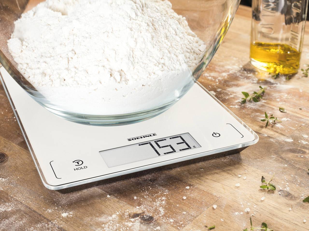 Une balance de cuisine numérique affiche 753 grammes. Un récipient en verre contenant de la farine est posé dessus. En arrière-plan, on peut voir de l'huile et des herbes.