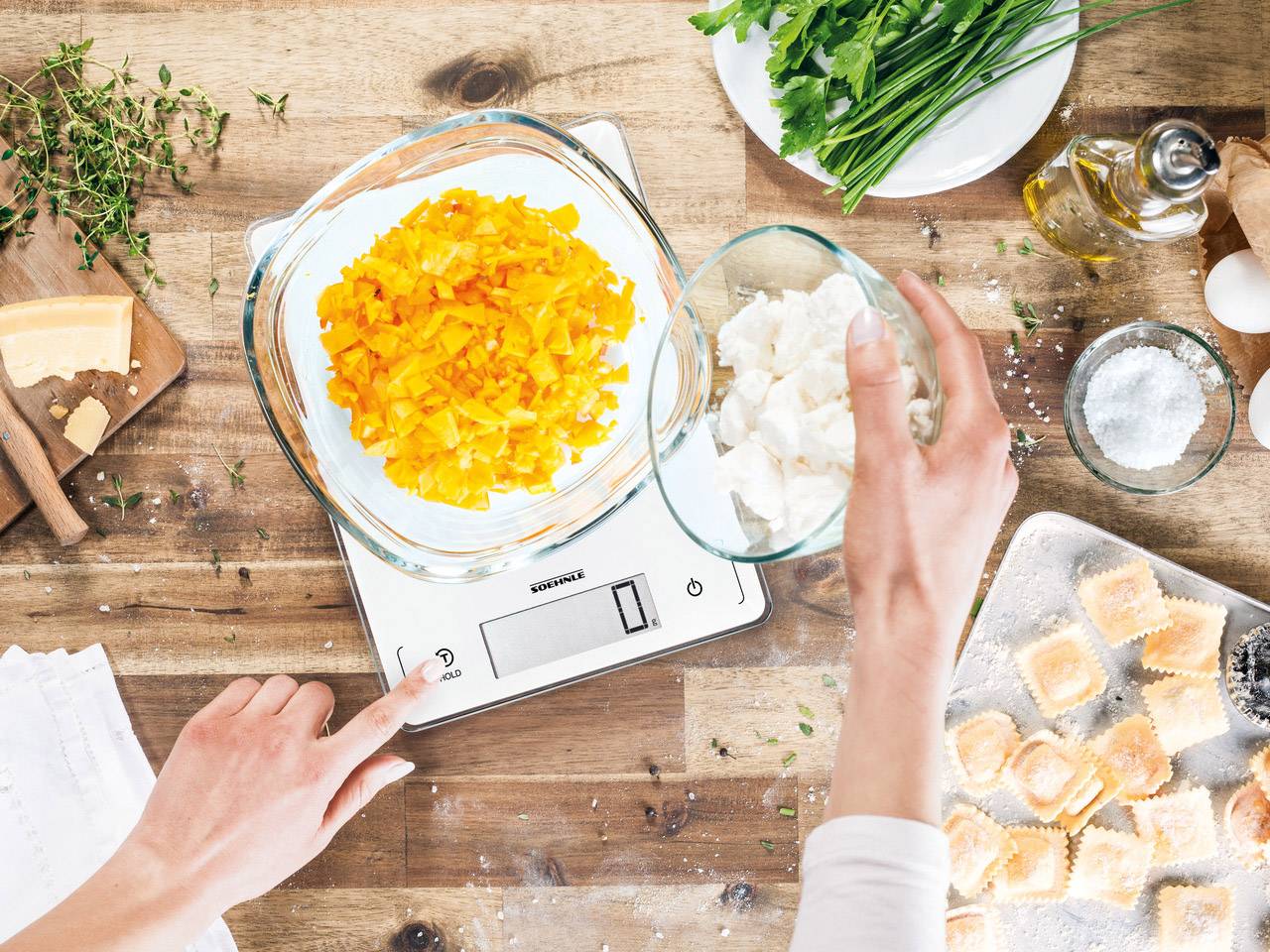Une personne pèse des dés de poivron jaune dans un bol sur une balance de cuisine numérique. En arrière-plan, des herbes fraîches et des raviolis.