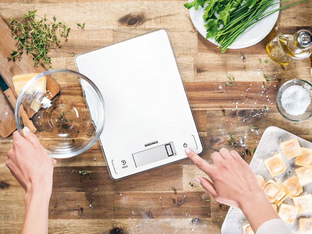 Une personne utilise une balance de cuisine numérique pour peser des ingrédients. Sur la table sont disposés des herbes, de l'huile et des raviolis crus.