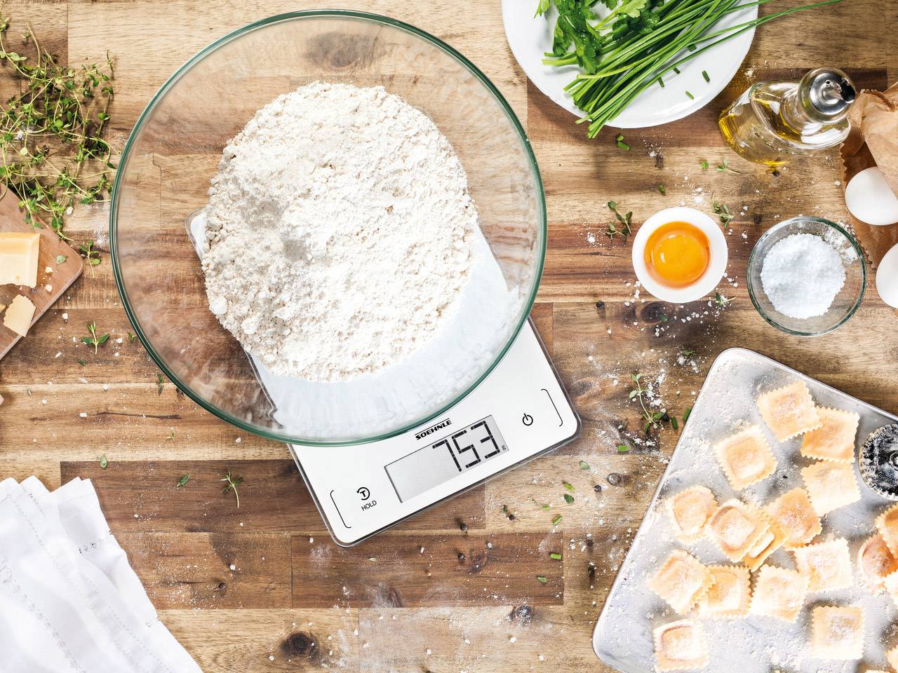 Bol de farine sur une balance de cuisine indiquant 753 g. Entouré d'herbes, d'œufs, d'huile et de morceaux de pâte sur une table en bois.