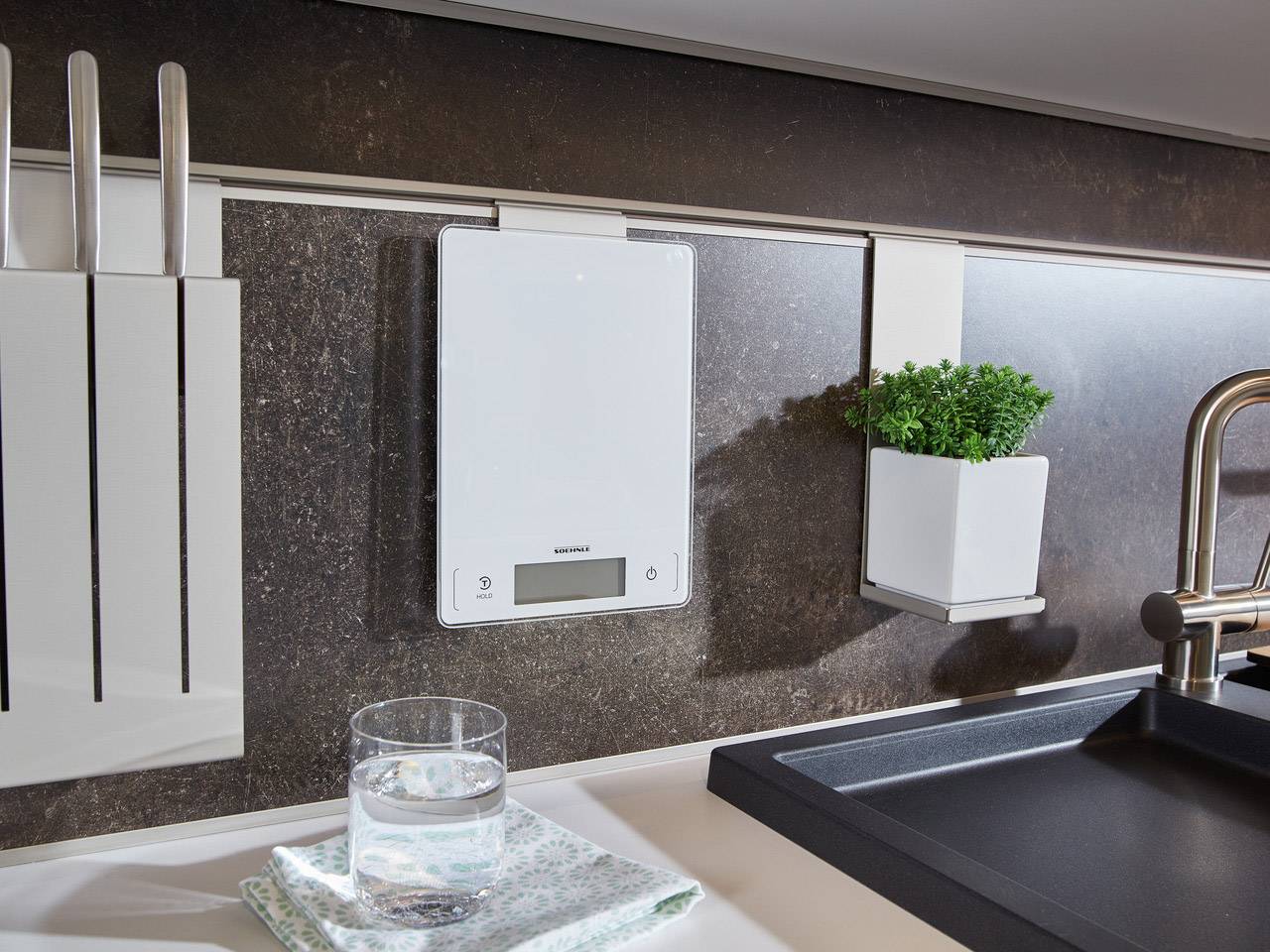 Une cuisine moderne avec une balance de cuisine numérique fixée au mur, à côté se trouve un pot avec des herbes fraîches. Un verre d'eau est posé sur le plan de travail.