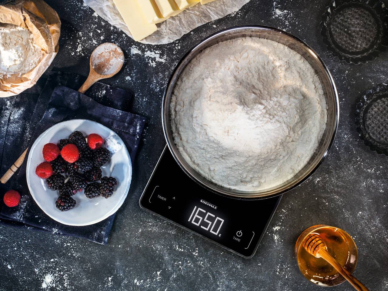 Bol de farine sur une balance indiquant 1650 grammes, à côté de baies, de beurre et de miel sur une table de cuisine sombre.