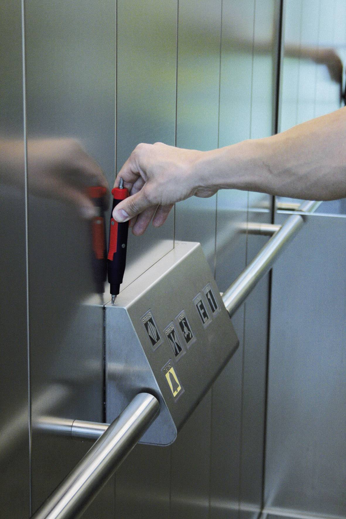 Un bras équipé d'un dispositif de test vérifie les touches d'un panneau de commande d'ascenseur. Les touches de sélection d'étage et d'appel d'urgence sont visibles.