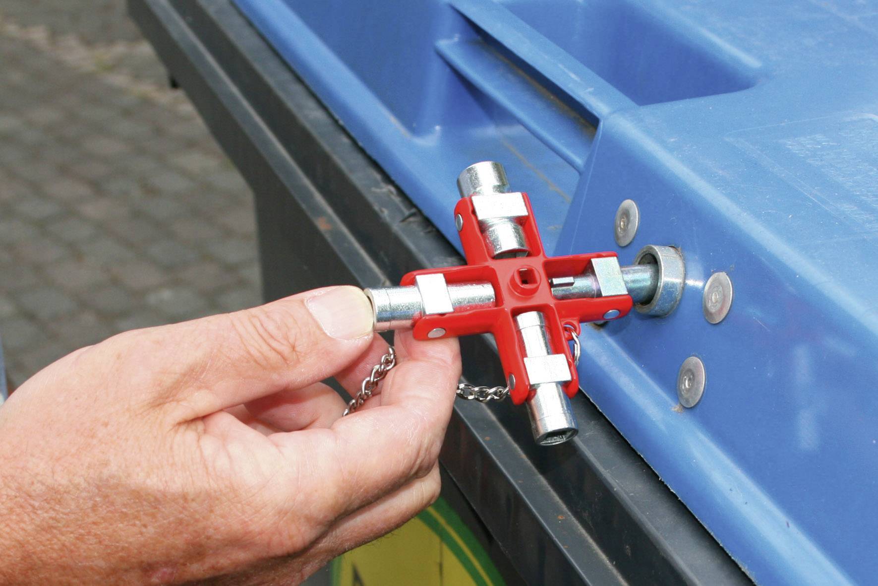 Une main tient une clé universelle rouge et déverrouille un mécanisme de fermeture sur un conteneur à déchets bleu.