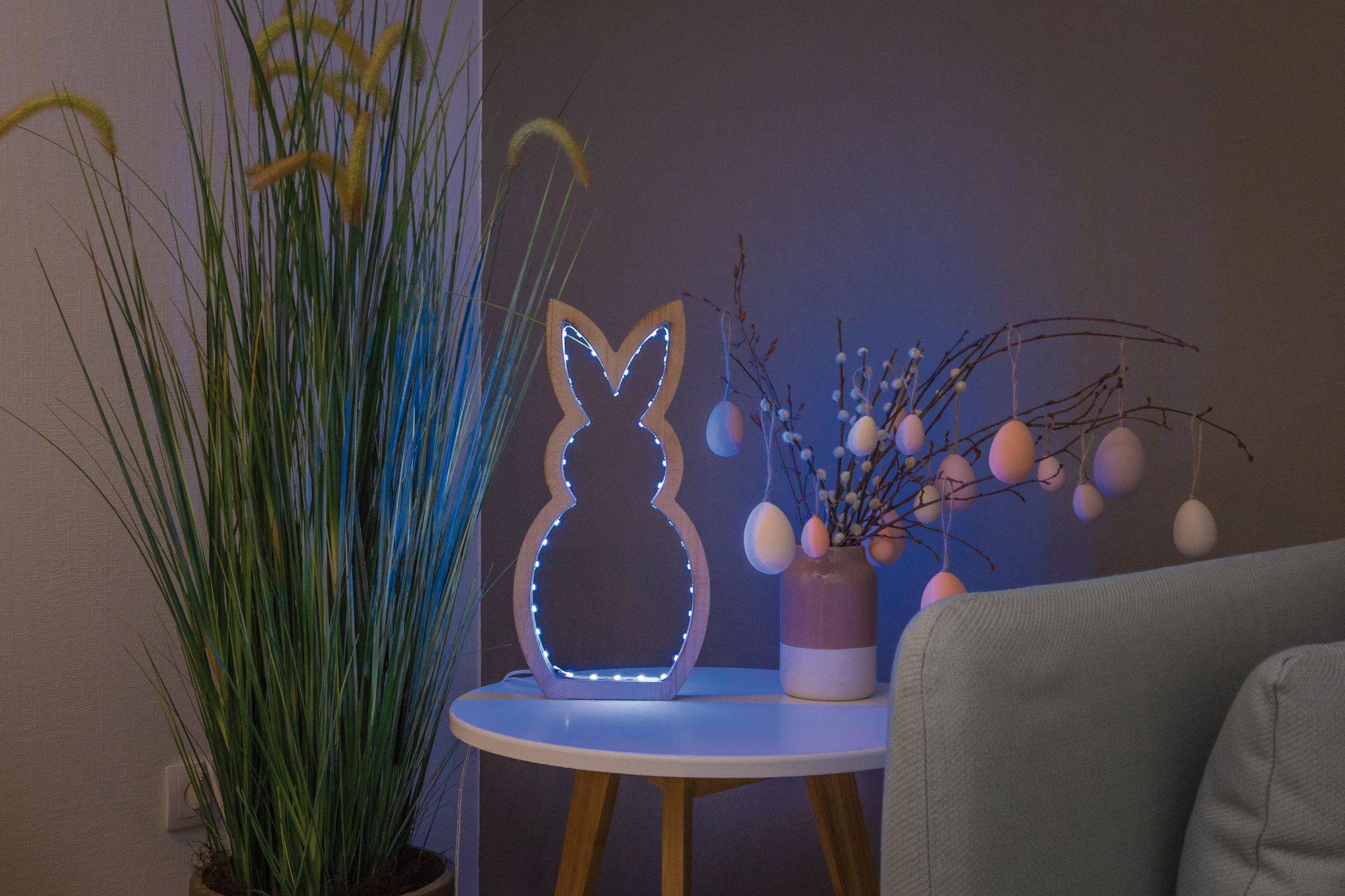 Une table décorative présente une figure de lapin lumineuse à côté de branches décorées d'œufs, entourée d'une plante d'intérieur haute.