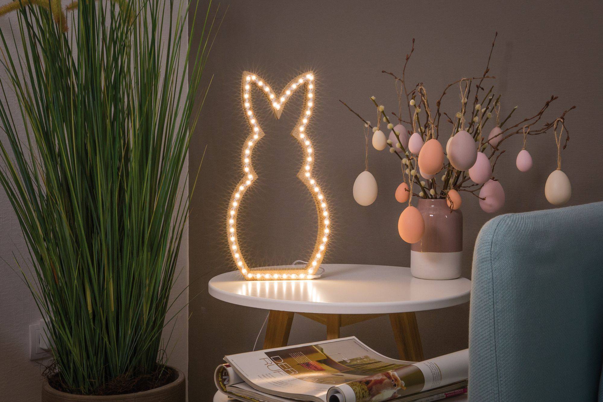 Décoration de Pâques illuminée en forme de lapin sur une table, à côté d'un bouquet avec des œufs de Pâques suspendus et une plante.