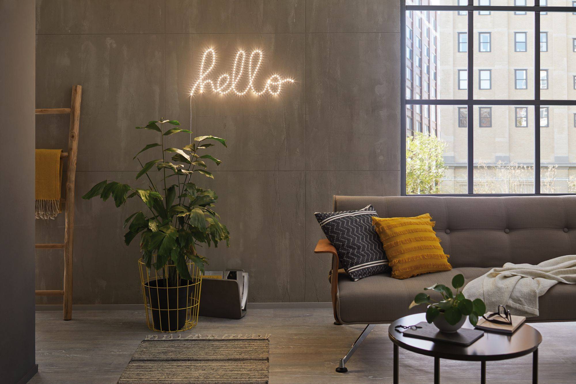 Salon avec un canapé gris, un coussin jaune, une plante dans un panier jaune et une décoration murale avec l'inscription lumineuse 'hello'. Fenêtre avec vue sur la ville.
