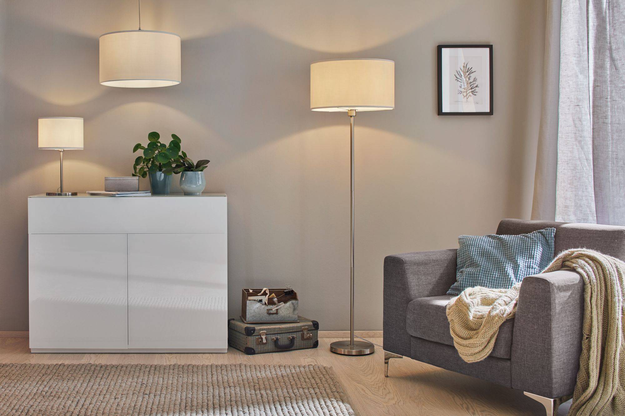 Un salon chaleureux avec un fauteuil gris, un buffet blanc, une lampe de table et une lampe sur pied. Des plantes et des décorations créent une ambiance accueillante.