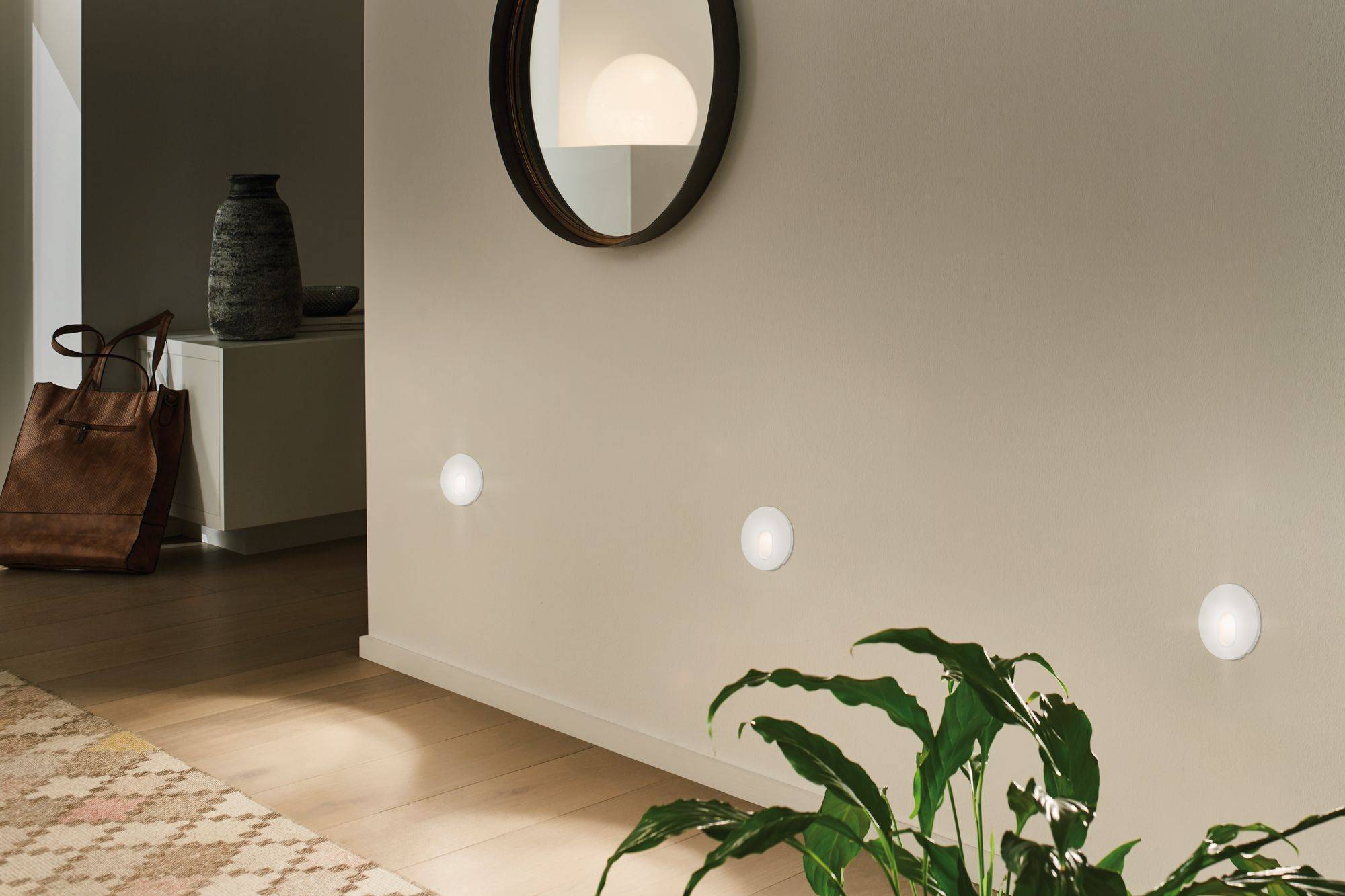 Un couloir avec un miroir accroché au mur, trois appliques murales rondes en dessous. À droite, en bas, une plante verte, à gauche un sac.