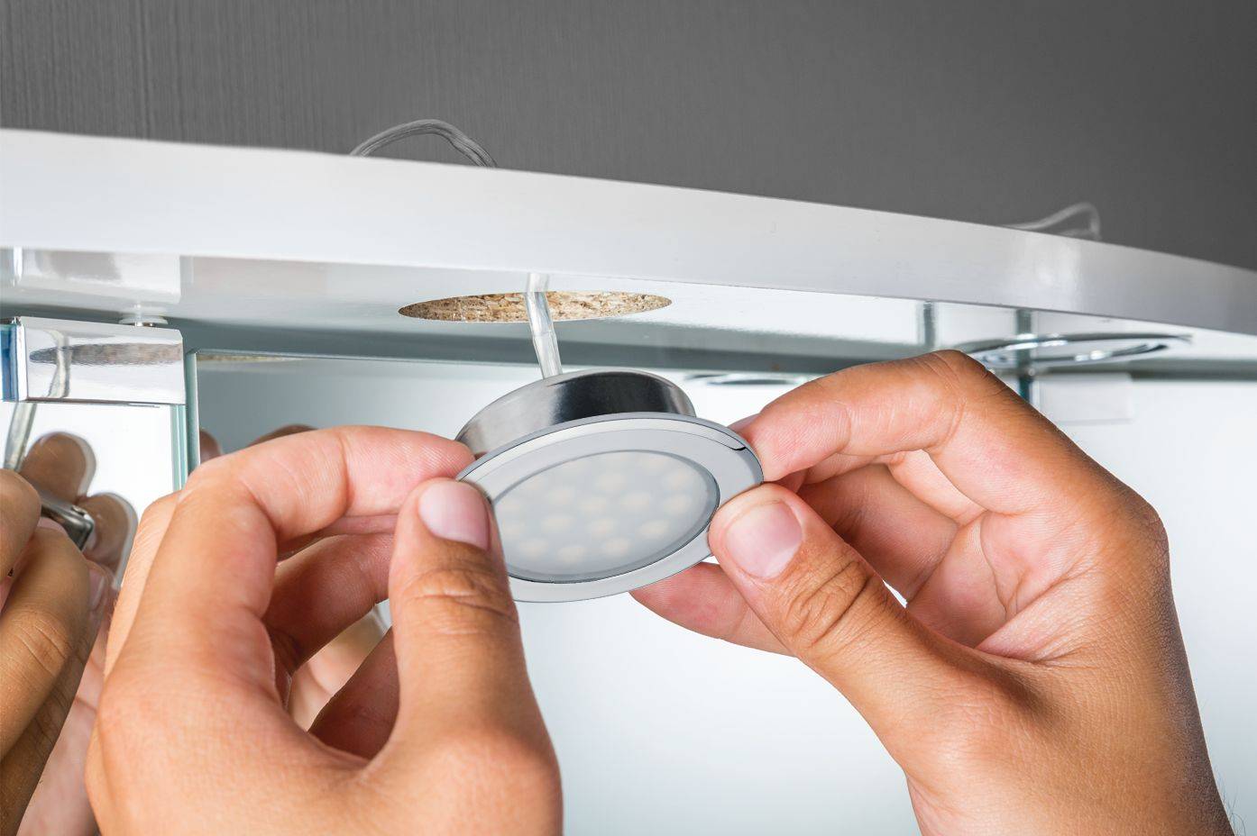 Une personne installe un spot LED rond et argenté au plafond près du bord d'un miroir. Les mains sont visibles près de la lumière.